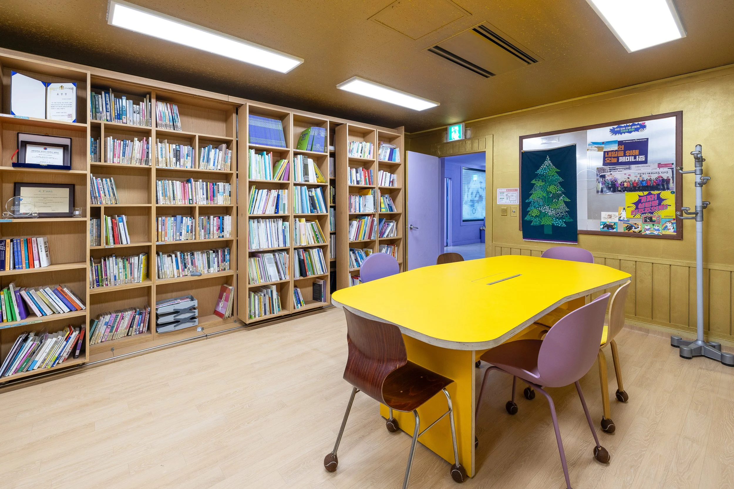 A room with wooden bookshelves filled with books, a yellow table, and chairs around it. The walls are decorated with posters and a bulletin board, and there is a coat rack. The room is lit with fluorescent ceiling lights.