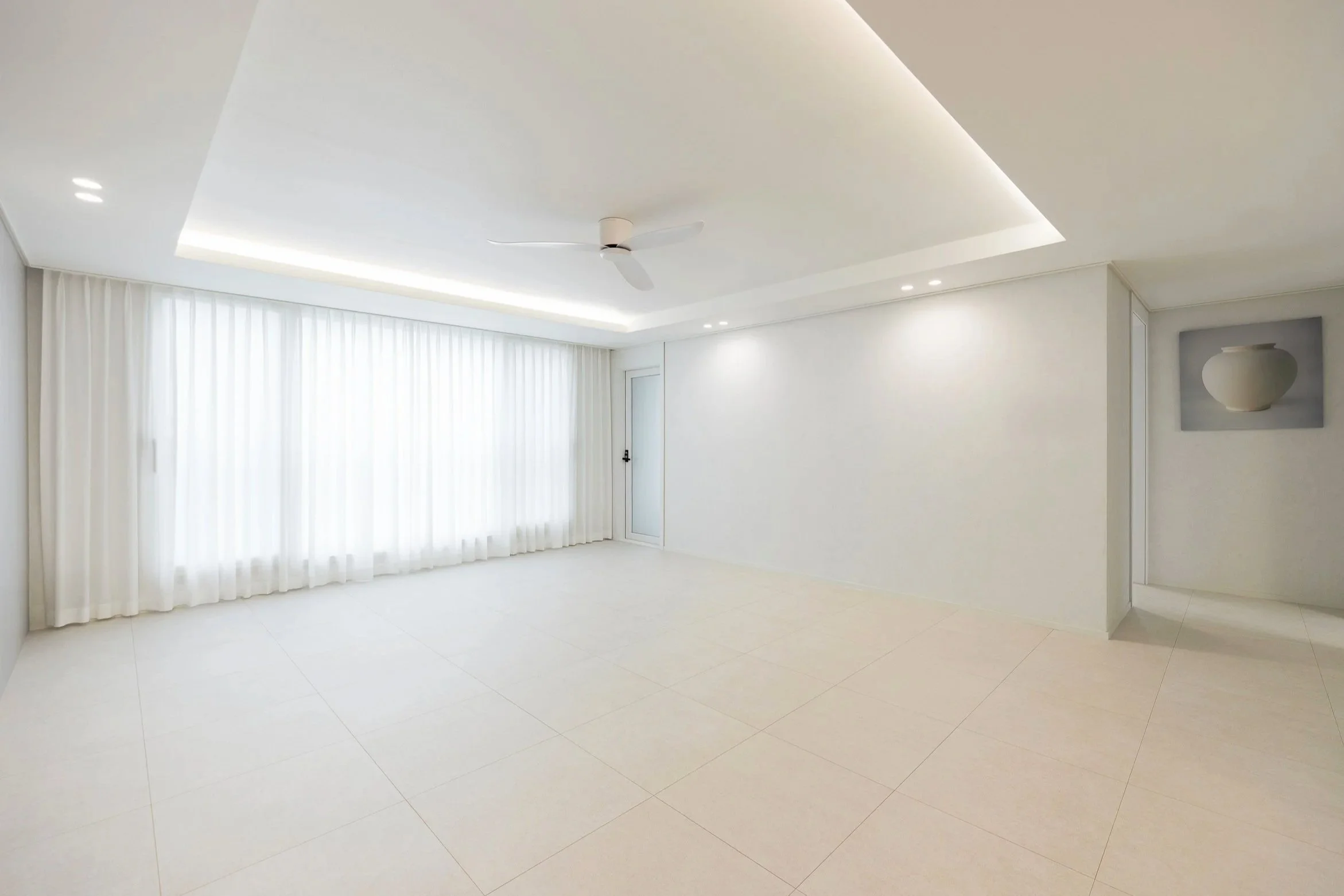 Bright, empty living room with white walls, large window with white curtains, ceiling fan, recessed lighting, and a single piece of artwork on the wall.
