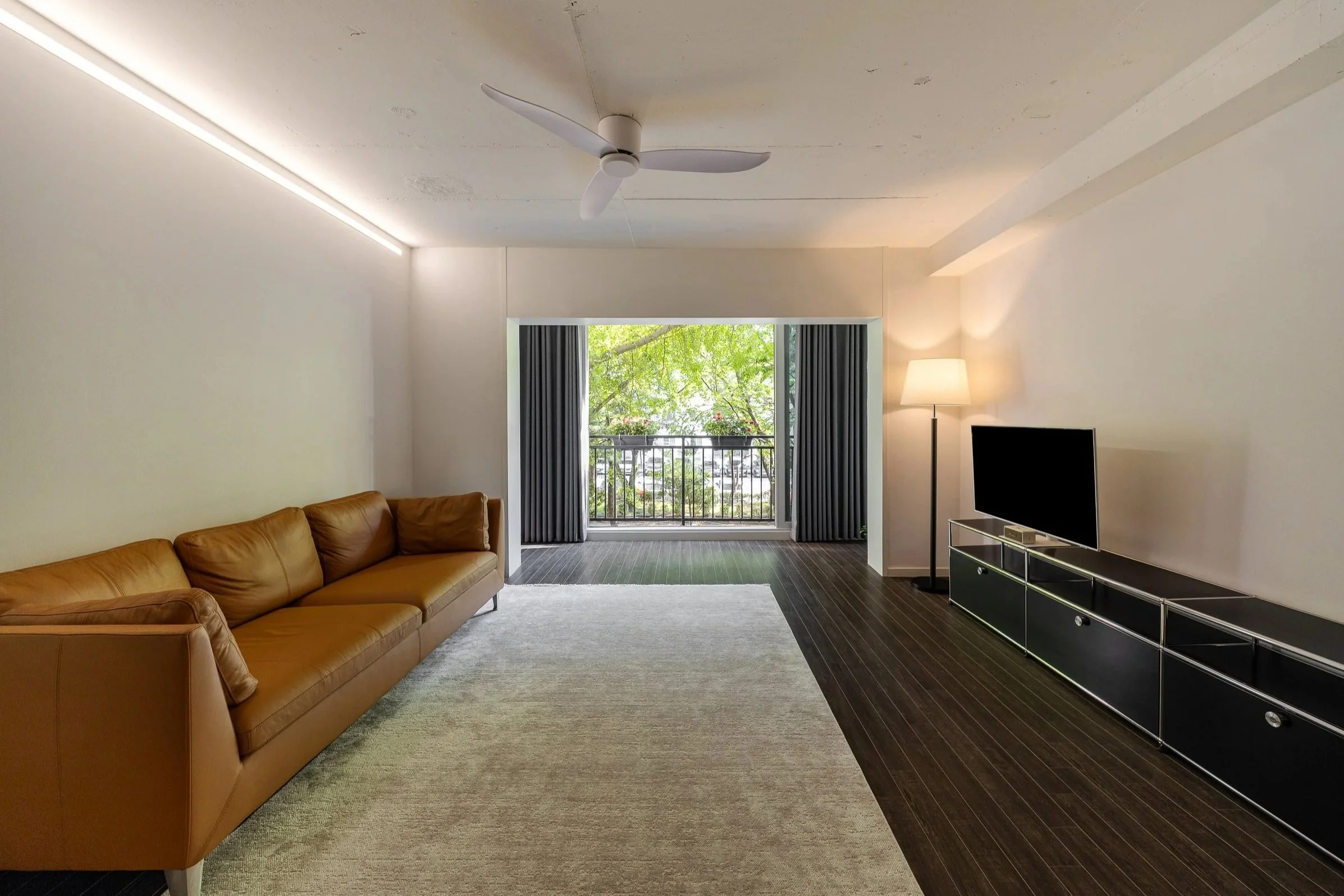 Living room with a large tan leather sofa, a black TV stand with drawers, a flat-screen TV, a floor lamp, a ceiling fan, and a balcony with trees visible outside, bright natural light illuminating the space.