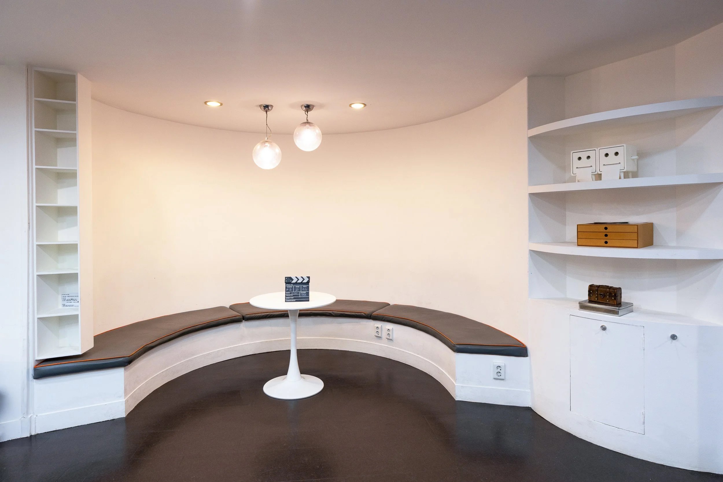 Empty room with curved bench seating, small round table, built-in white shelves, and pendant lighting