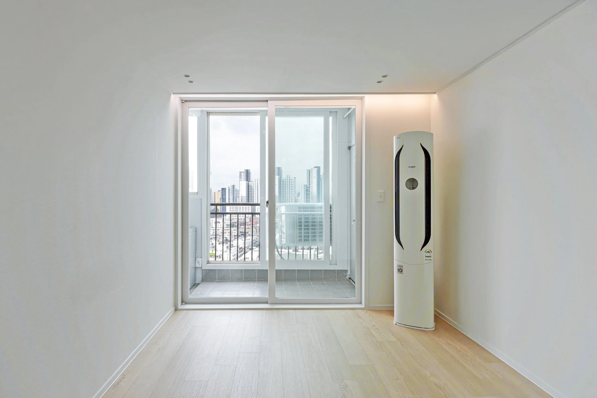 Empty room with wooden floor, white walls, sliding glass door leading to a balcony, and view of city skyscrapers