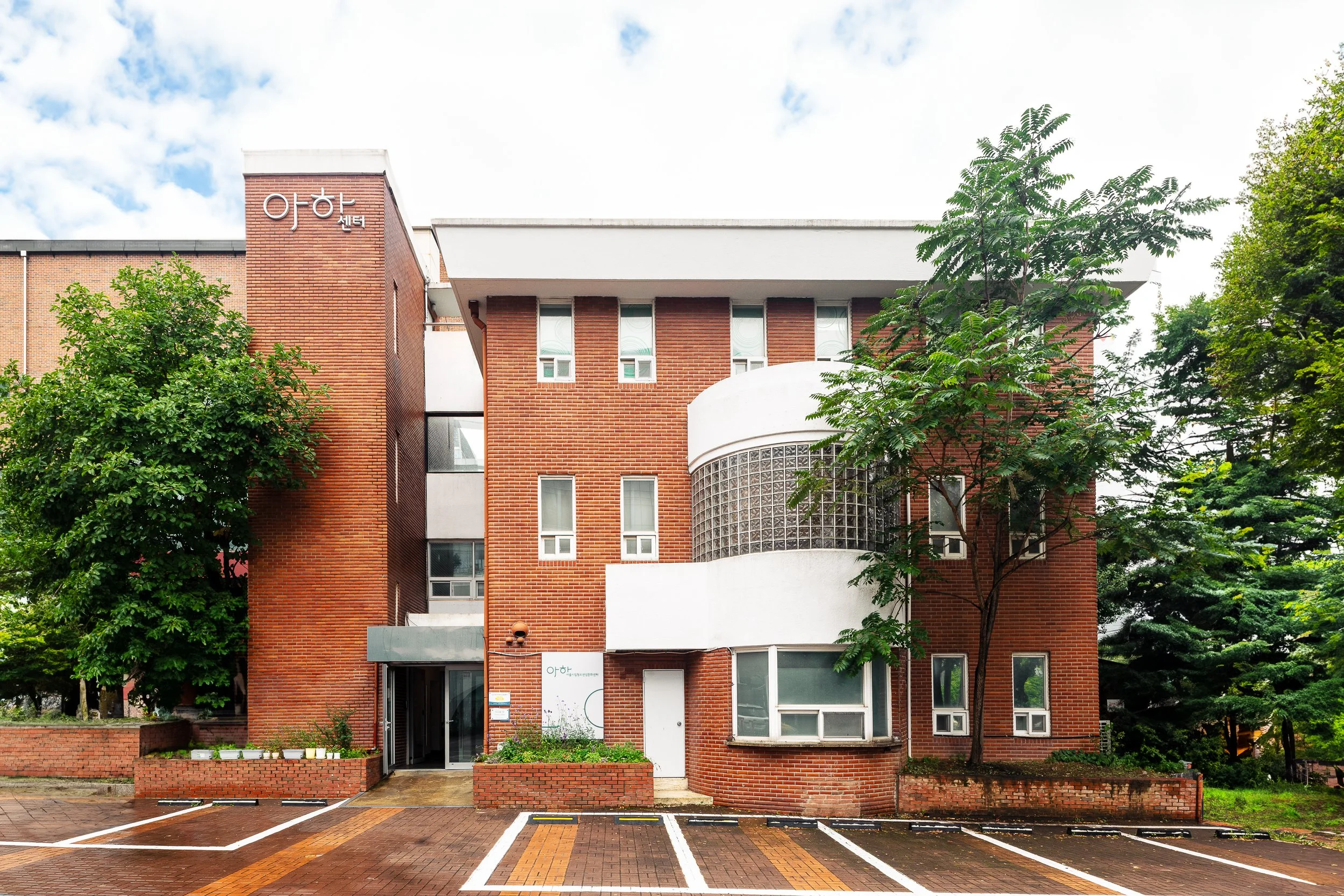 A multi-story brick building with white accents, surrounded by green trees, with a parking area in front and a cloudy sky above.