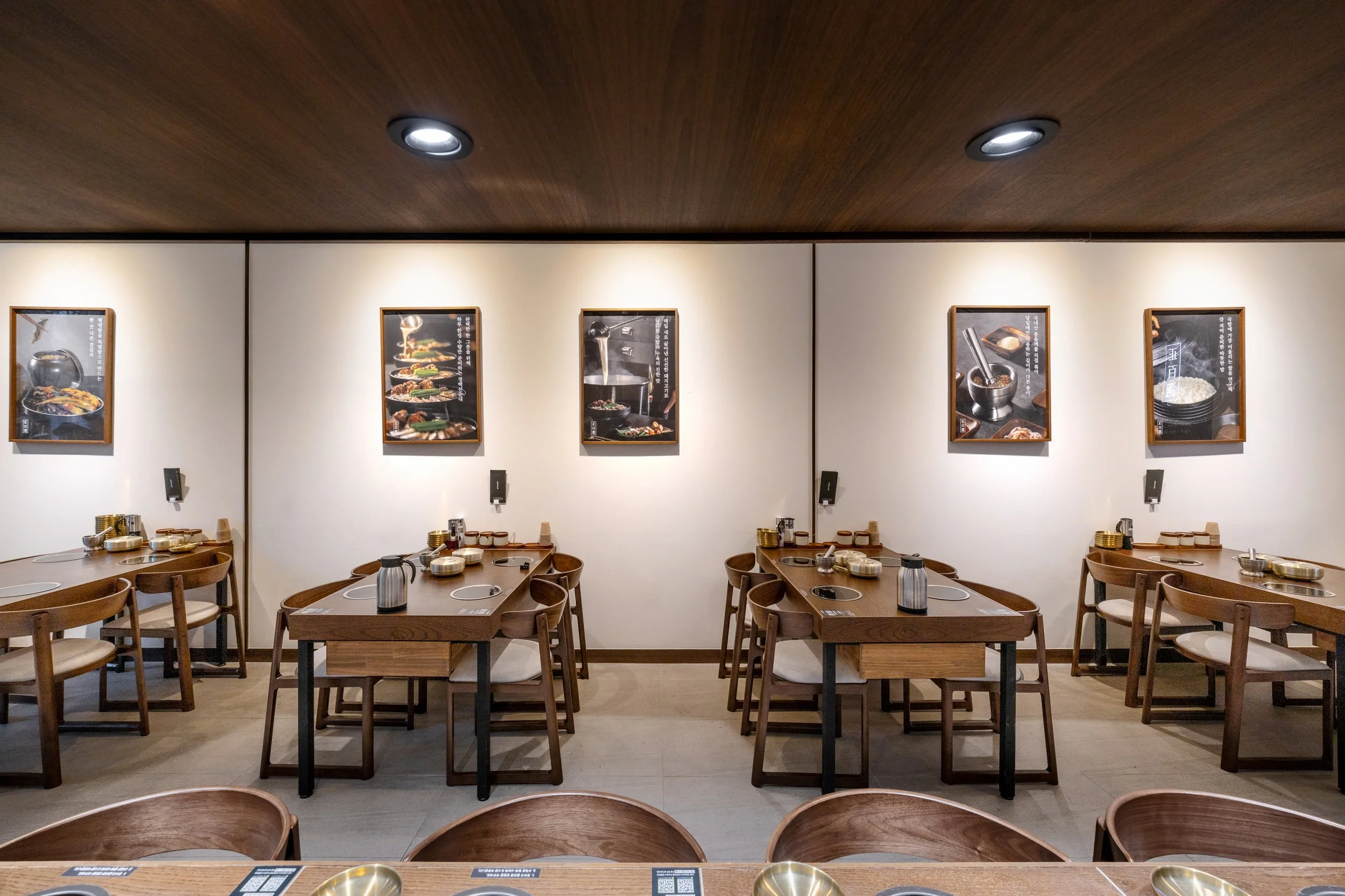 Empty Japanese restaurant dining area with wooden tables and chairs, wall art depicting food and cooking, and ceiling spotlights.