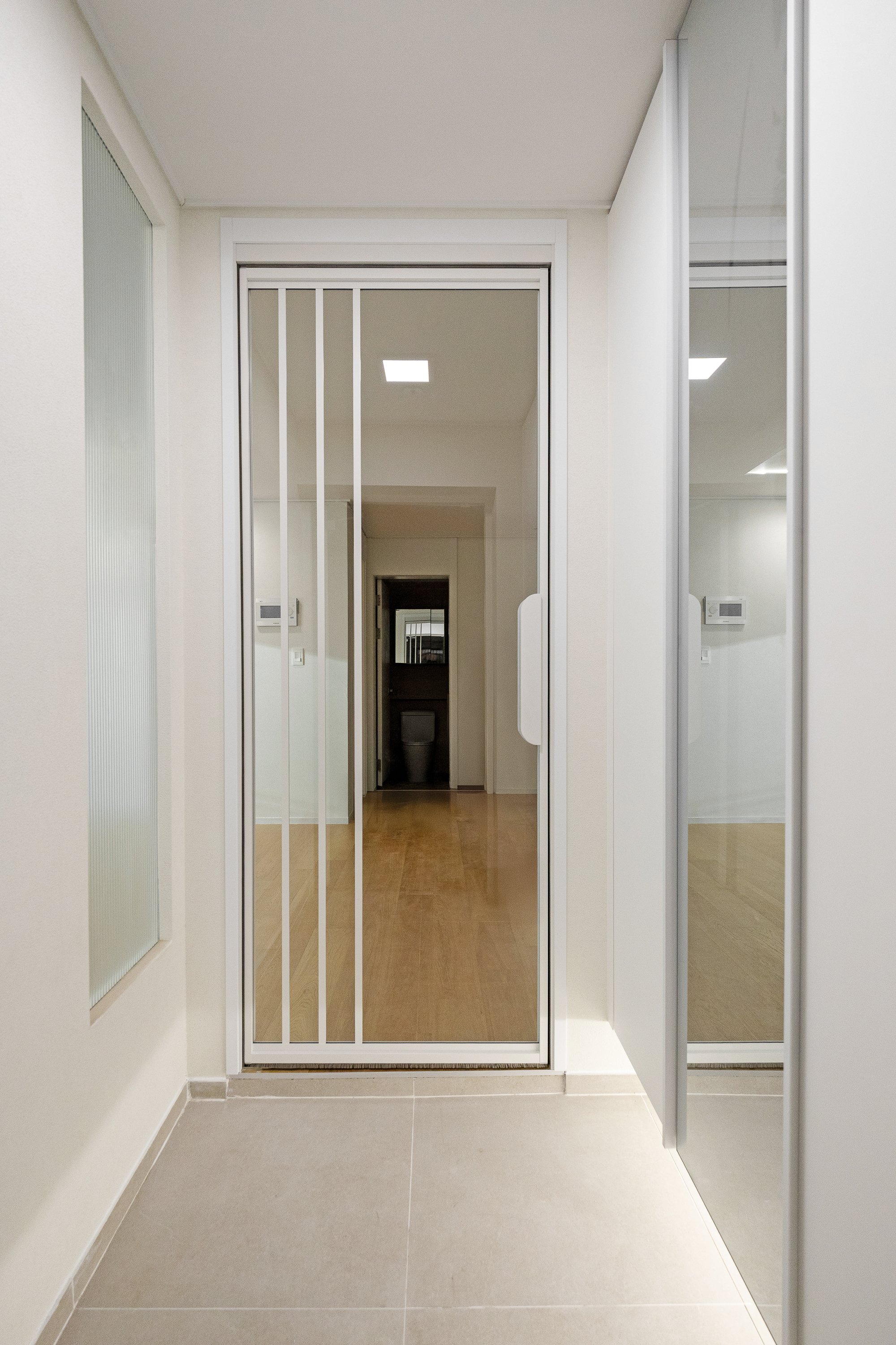 Interior view of a modern apartment entrance with a glass and metal sliding door, tiled floor, and visible hallway leading to a bathroom.