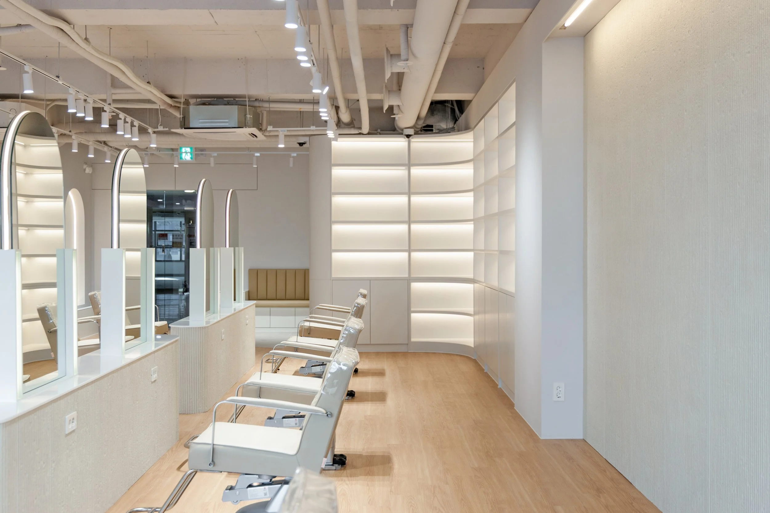 Empty modern salon with white chairs, light wood flooring, and illuminated wall shelves.
