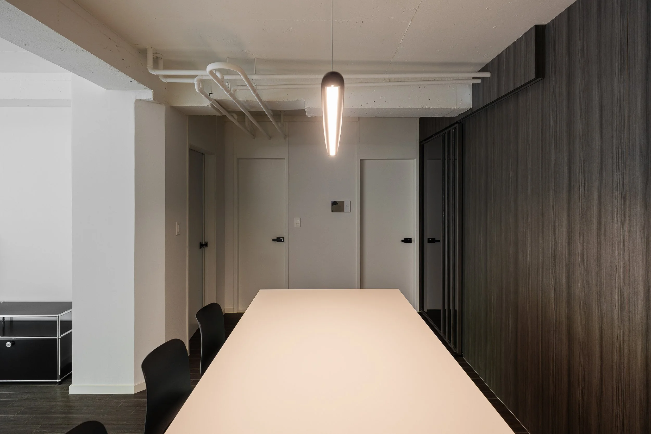 Modern conference room with a long white table, black chairs, dark wood accent wall, white ceiling with visible pipework, and a hanging light fixture.