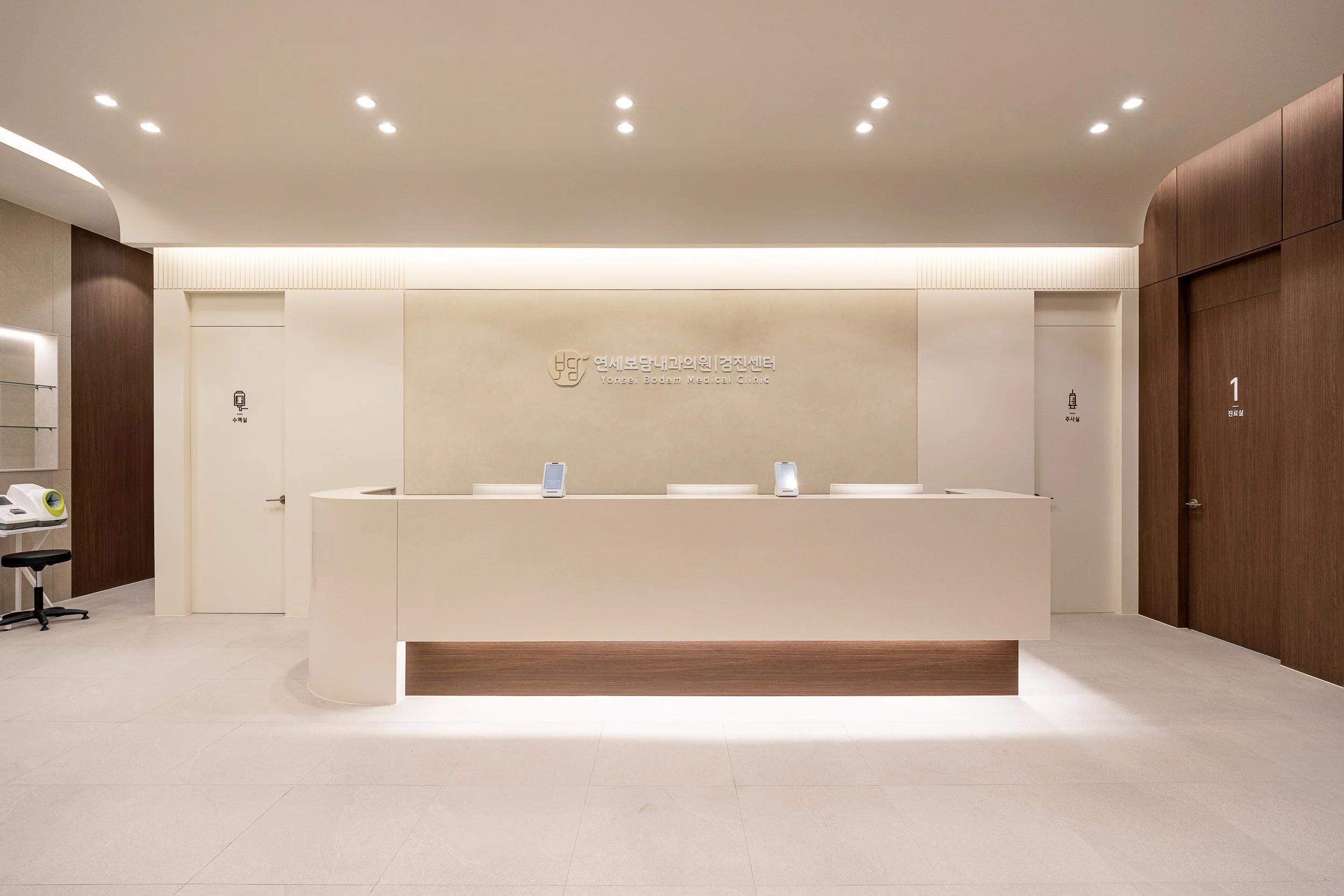 Reception area of Yonsei Bodam Medical Clinic, featuring a white curved counter with digital devices, beige walls, and wooden accents. Doors marked for restrooms and room 1 are visible.