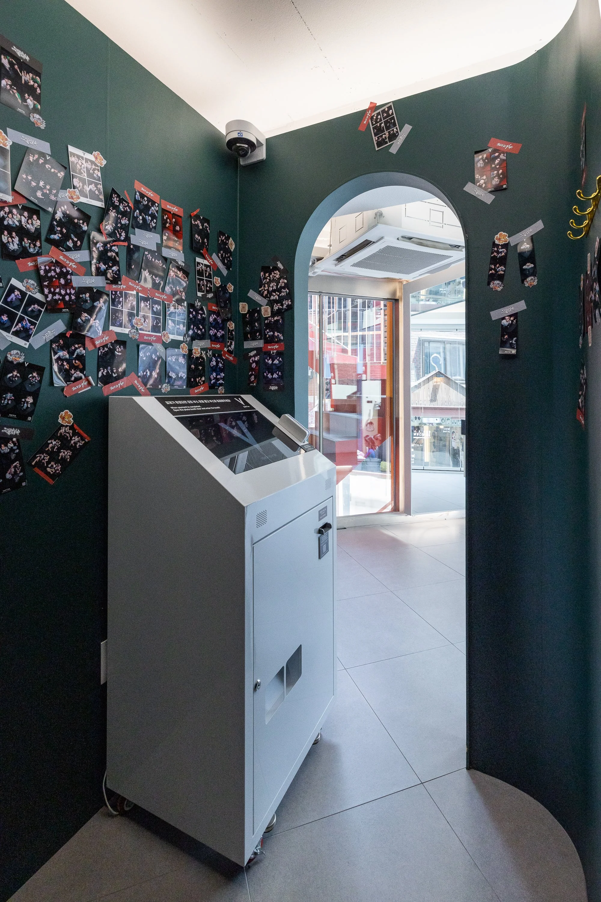 Dark green walls decorated with numerous photo prints and small decorations, with a white and gray kiosk or vending machine in the center. A glass door leading outside shows outdoor seating and a street scene.