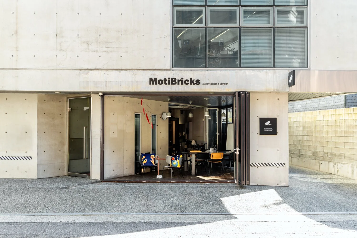 Exterior of a modern building with a cafe named MotiBricks, featuring an outdoor seating area with colorful chairs. The building has concrete walls, large windows on the upper floor, and a sign with a creative design logo.