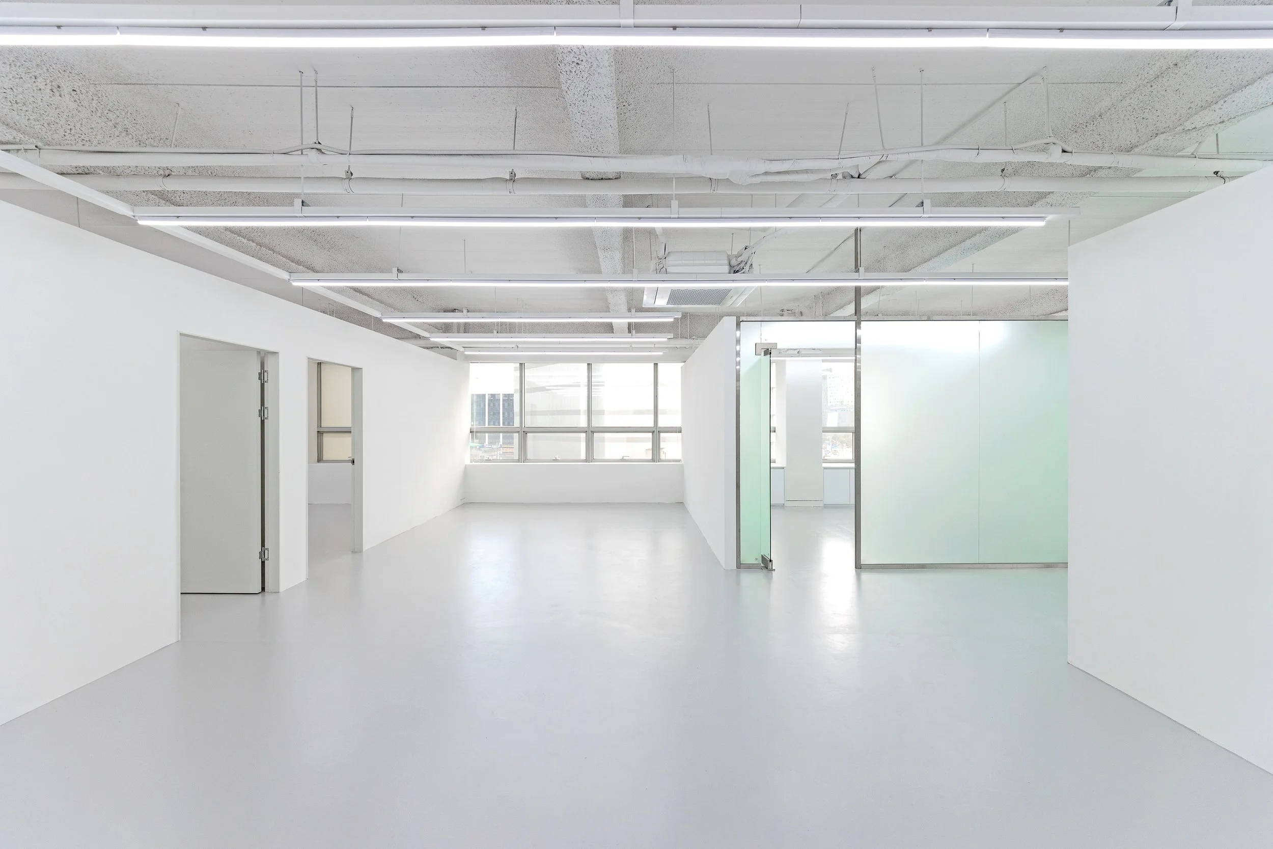 Bright, minimalist office space with white walls, large windows, and an open layout with glass partitions.