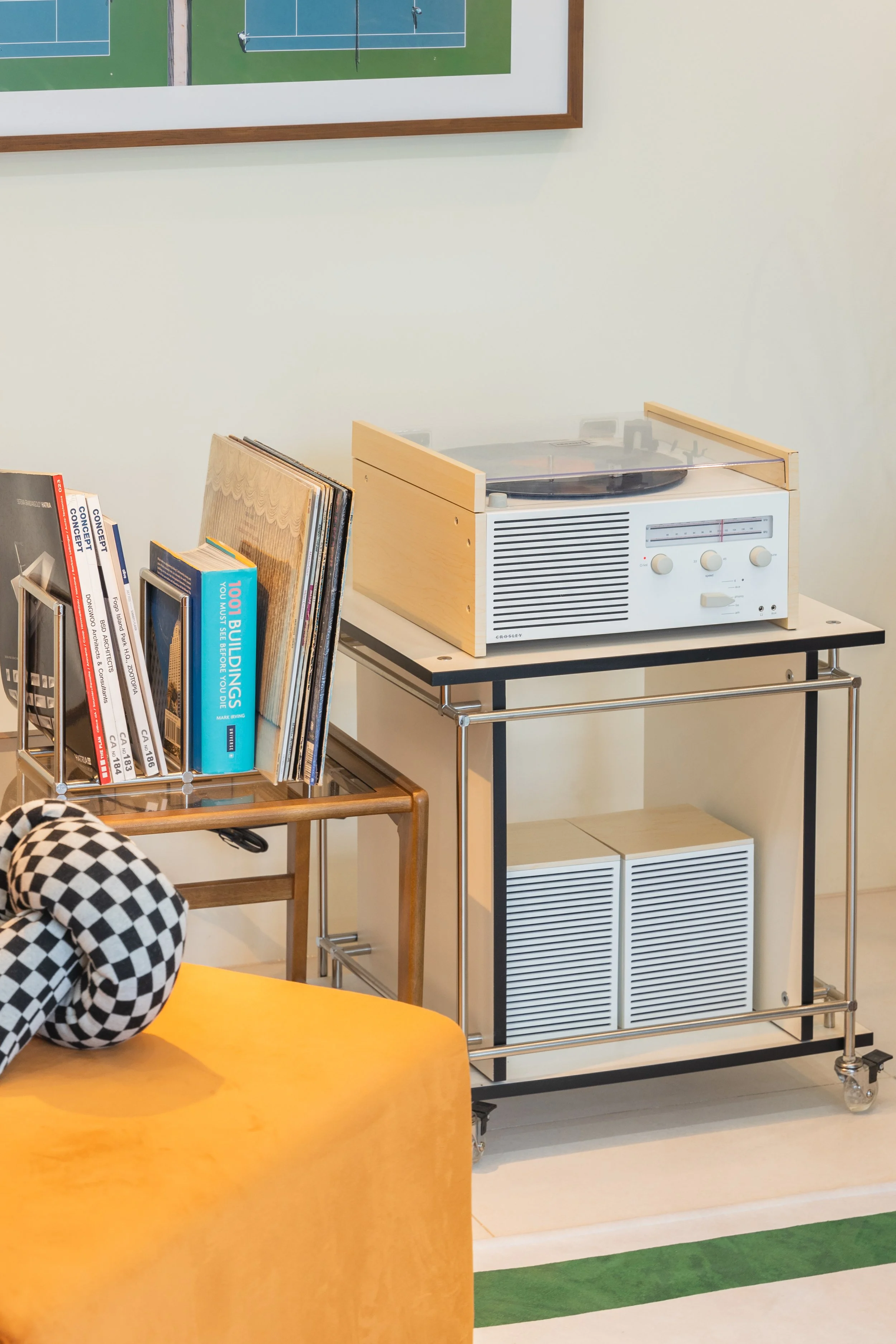 A vintage turntable on a black metal and wood cart, a bookshelf with books, and part of a yellow sofa with a checkered black and white pillow.