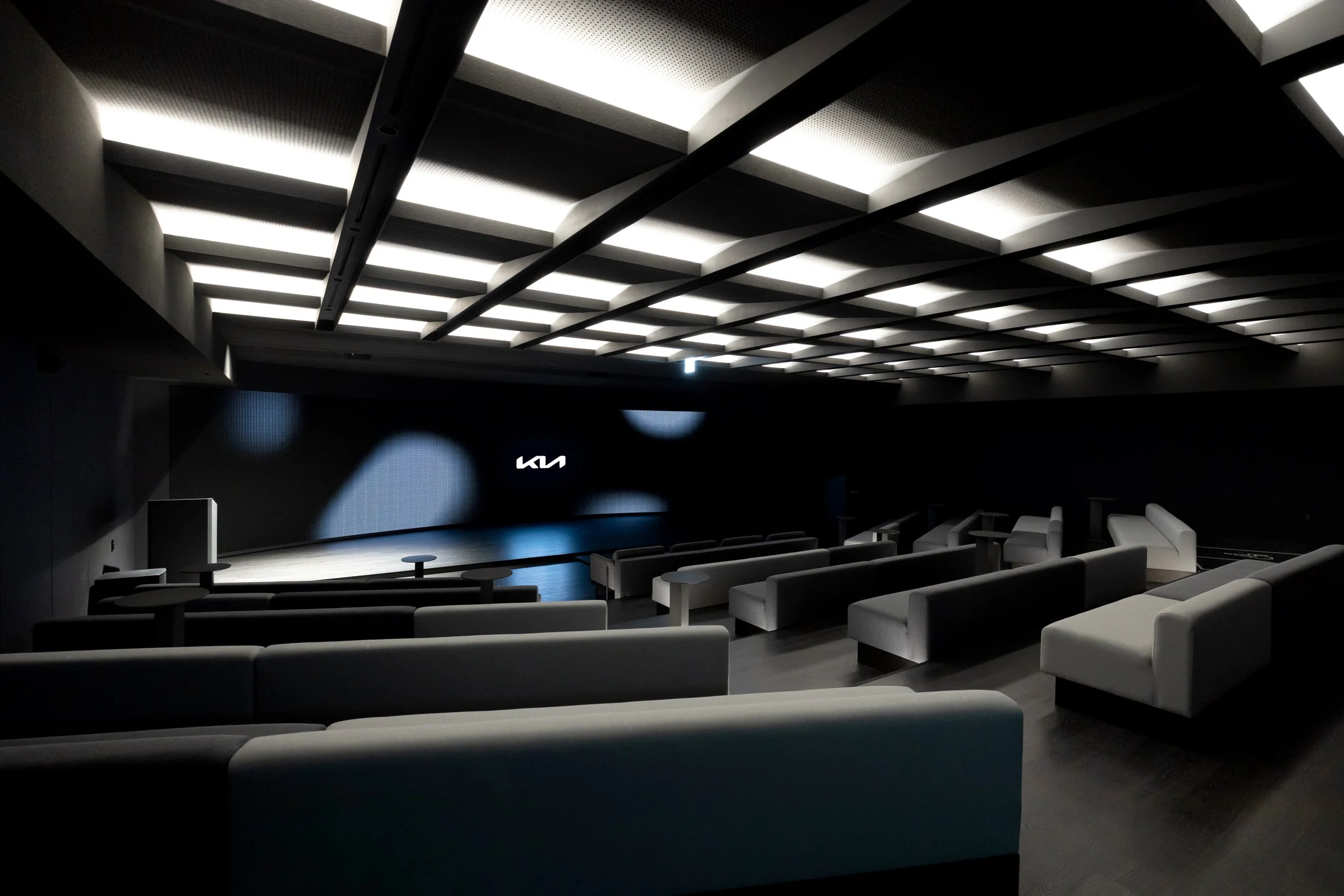 Dark, empty modern auditorium or movie theater with black walls, white ceiling panels with lighting, and white seating arranged in rows facing a large screen with the Kia logo.