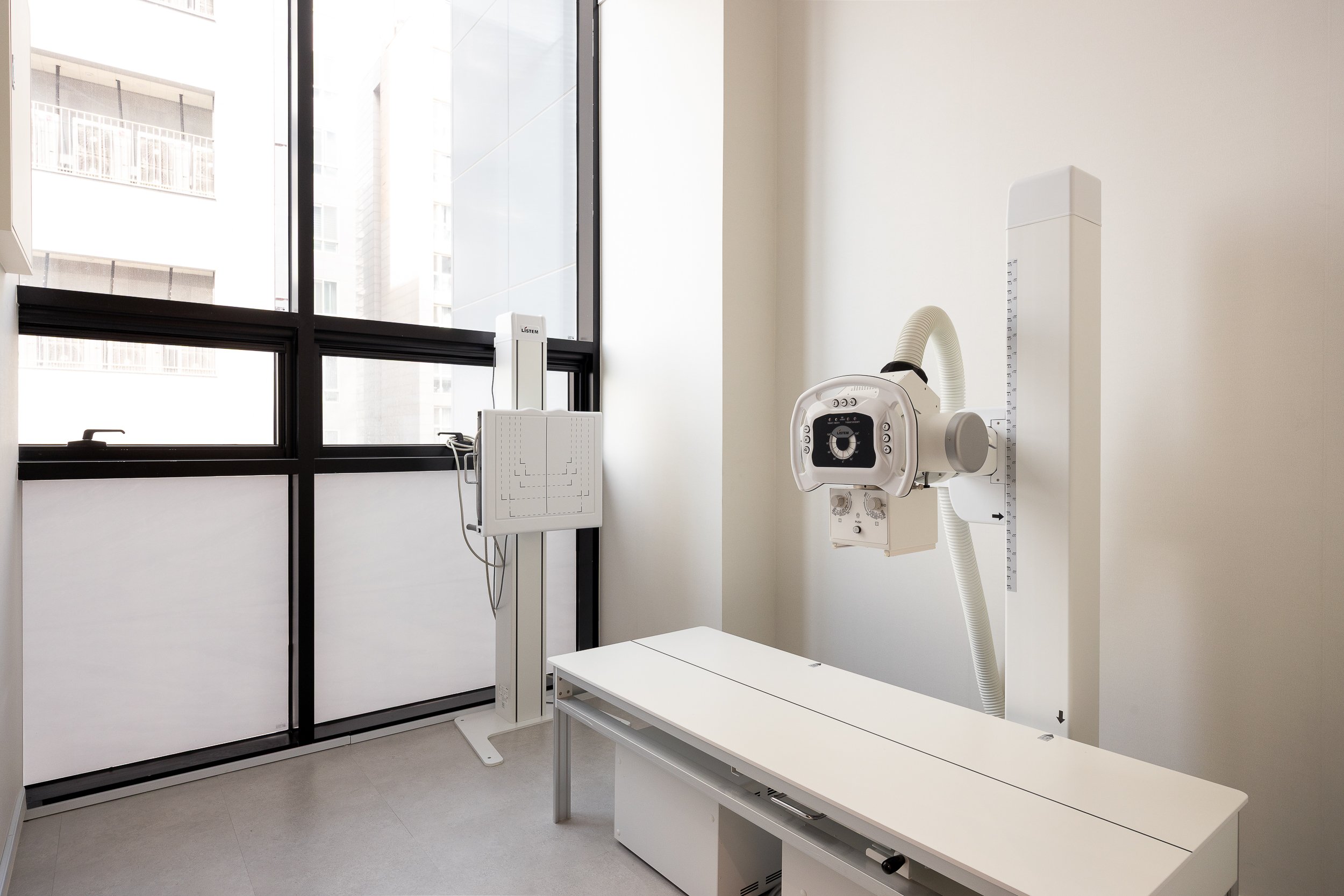 An empty medical X-ray room with a white X-ray machine mounted on the wall and a patient examination table.