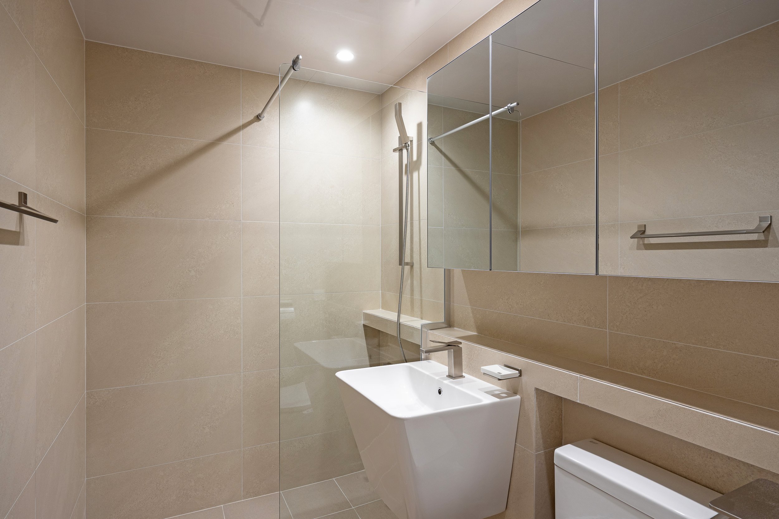 A modern bathroom with beige tiled walls and floor. It features a white, vessel sink with a chrome faucet, a large mirror above the sink, a toilet to the right, and a walk-in shower area separated by a glass partition, with a showerhead and a towel b