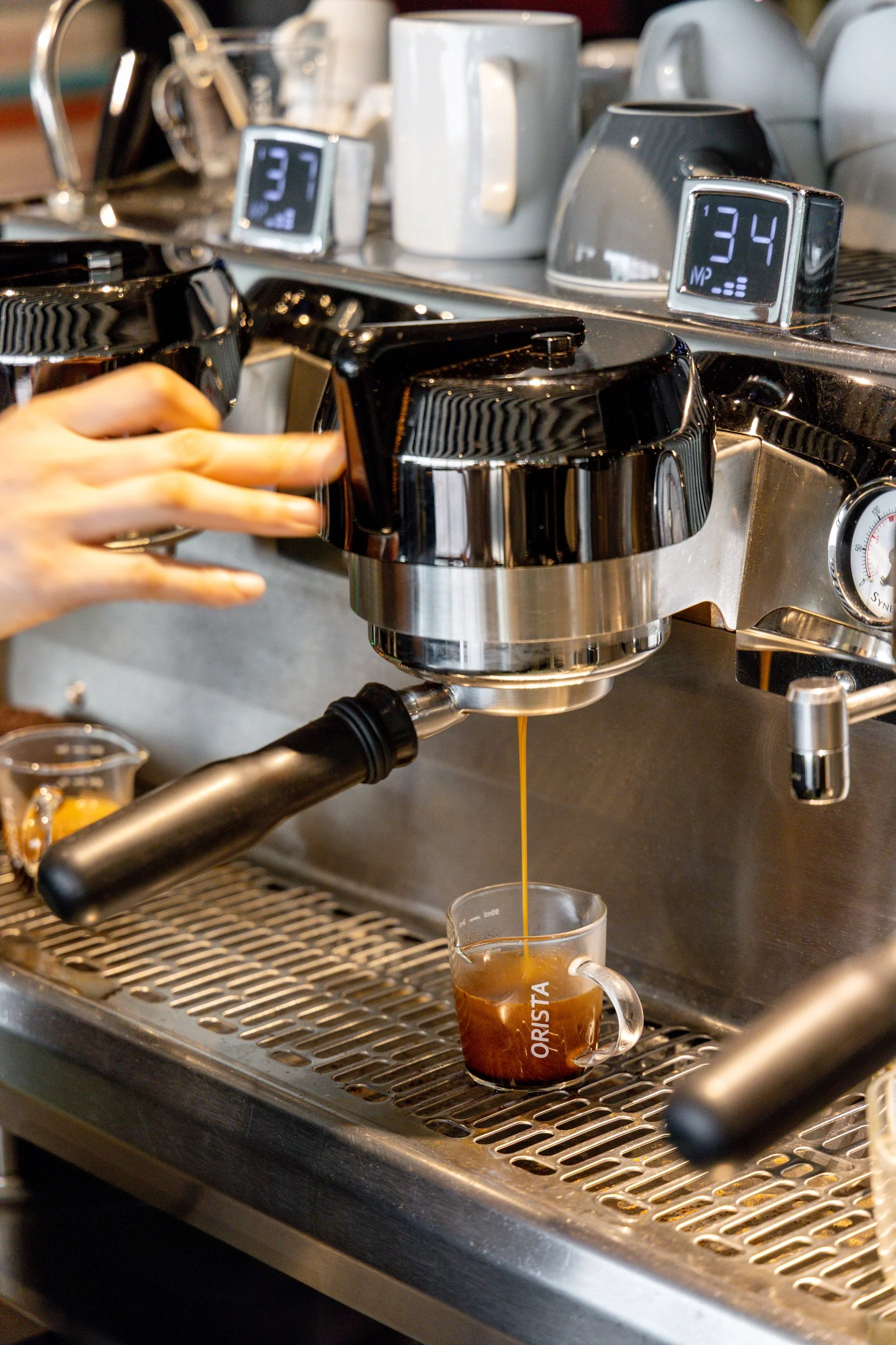 Close-up of a barista preparing an espresso with a professional espresso machine, with a small glass cup labeled ORISTA underneath, and various coffee mugs and digital screens in the background.