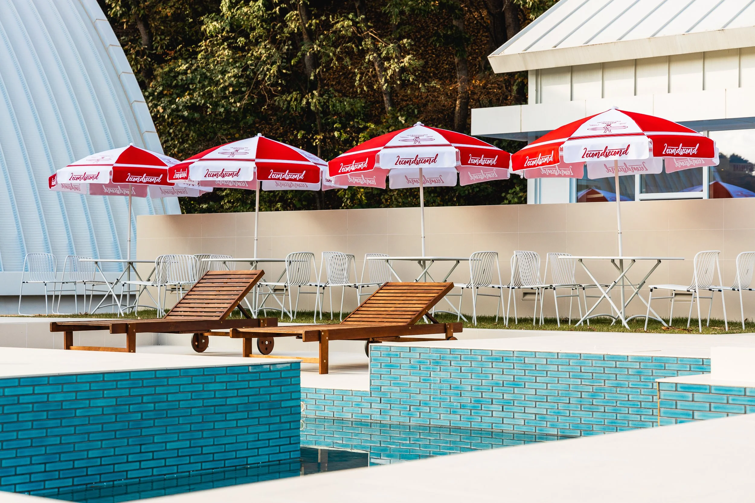 Empty poolside area with wooden lounge chairs, white metal chairs, and red and white umbrellas at a sunny outdoor poolside area.
