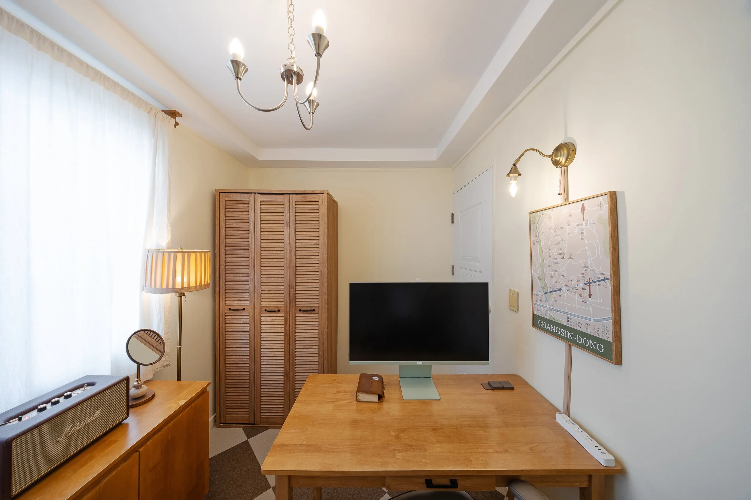 A cozy home office with a wooden desk holding a computer monitor, a notebook, and a mouse, near a map on the wall and a shelf with a voice amplifier, accompanied by a standing lamp, a window with white curtains, a wooden wardrobe, and a ceiling chand