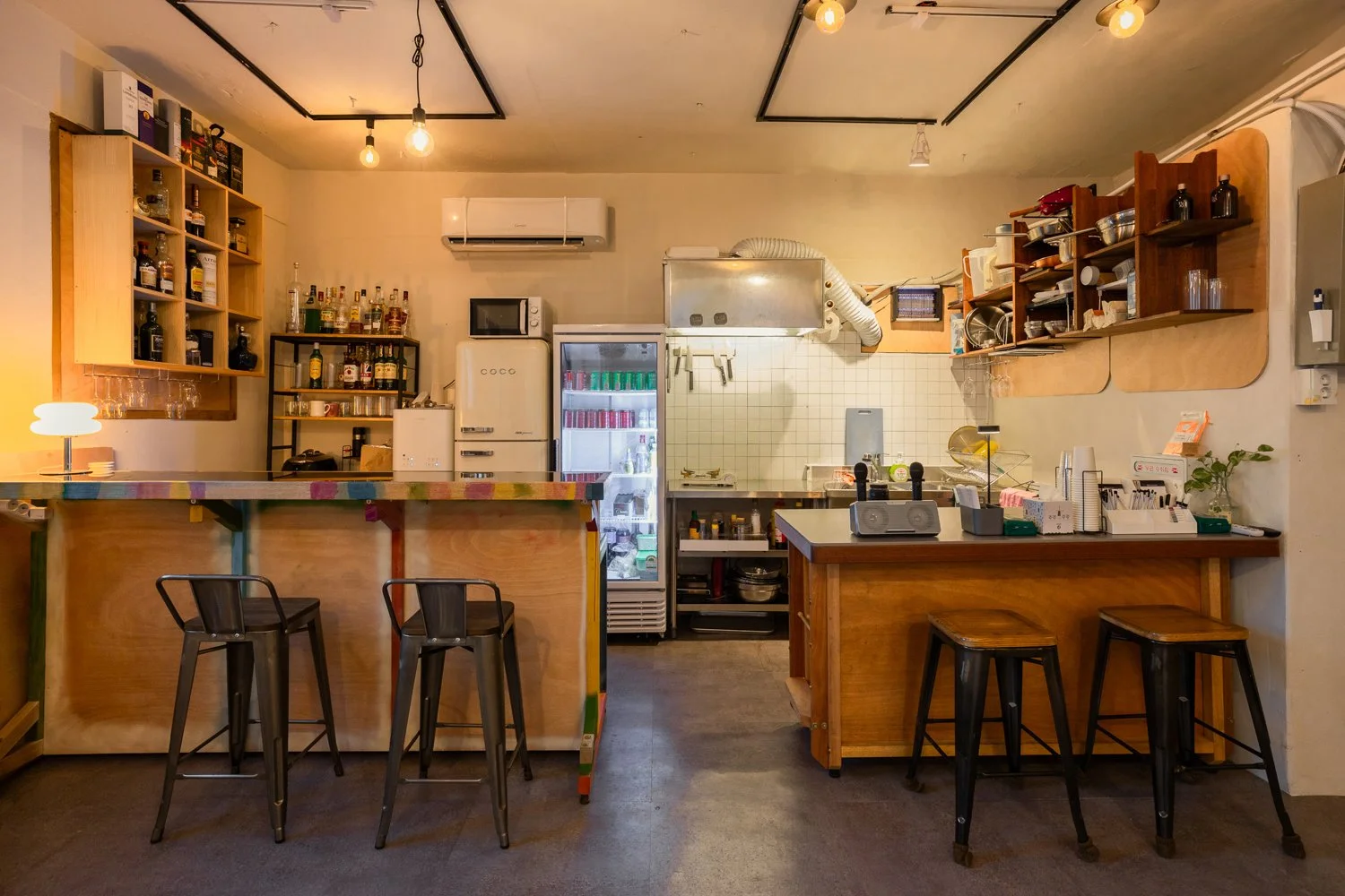 Interior of a small cafe with a wooden counter, bar stools, shelves with bottles and glasses, a refrigerator with drinks, and a kitchen area with utensils and appliances.