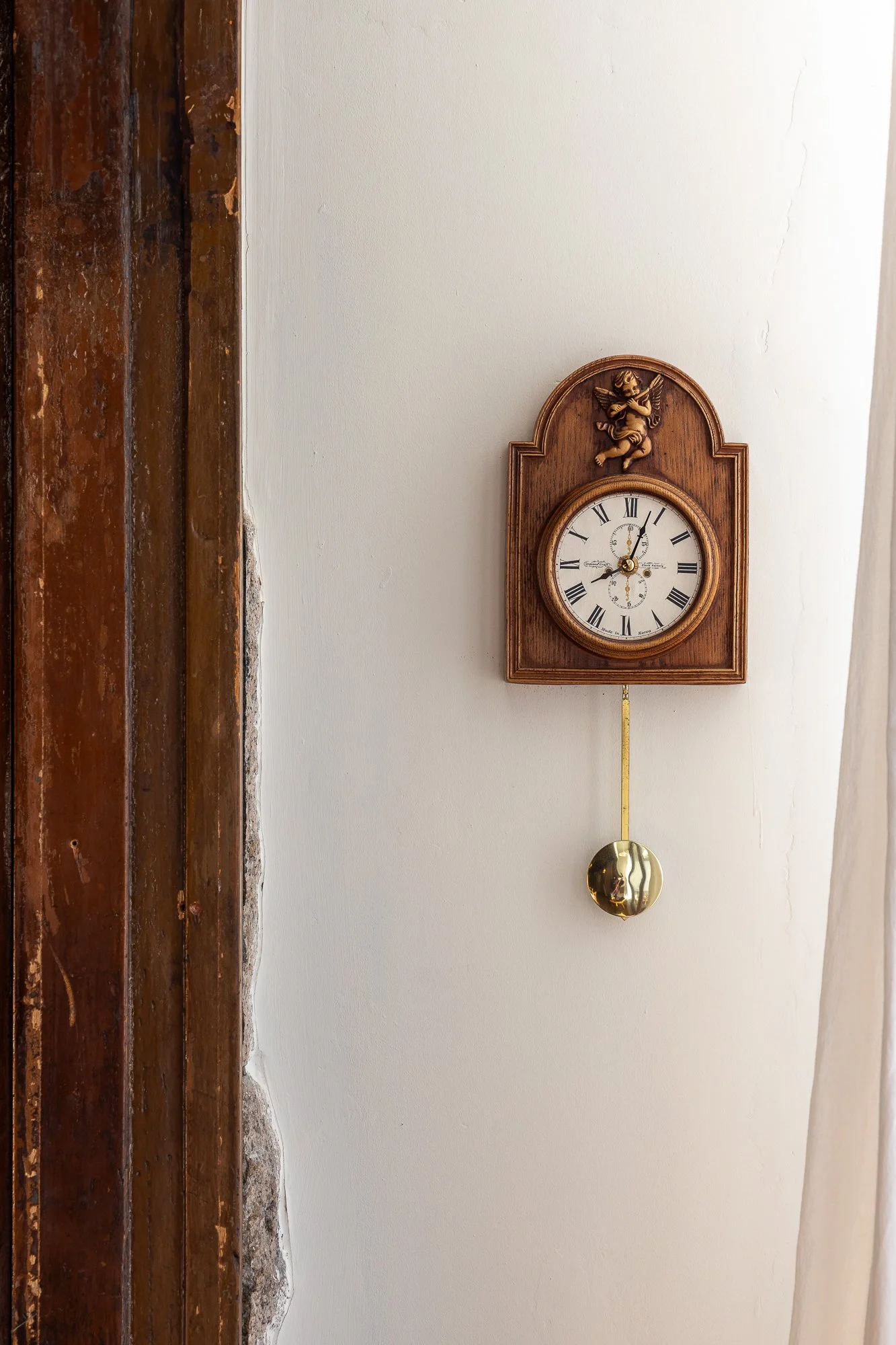 A wooden wall clock with a cherub design at the top, featuring Roman numerals and clock hands, and a gold pendulum hanging below.