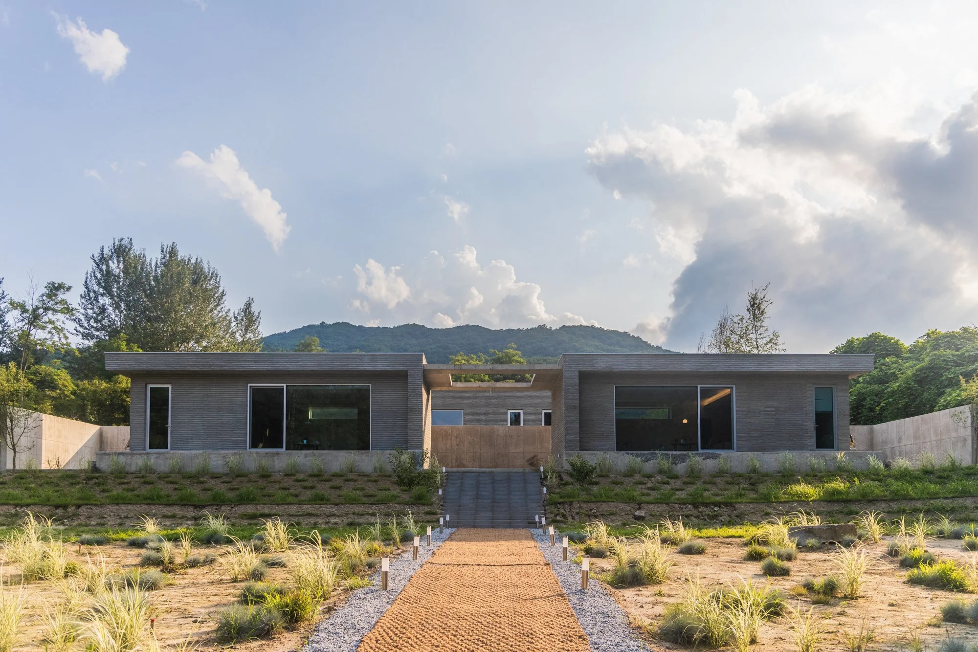 Modern house with large glass windows, situated on a landscaped lot with plants, leading to a mountain in the background under a partly cloudy sky.