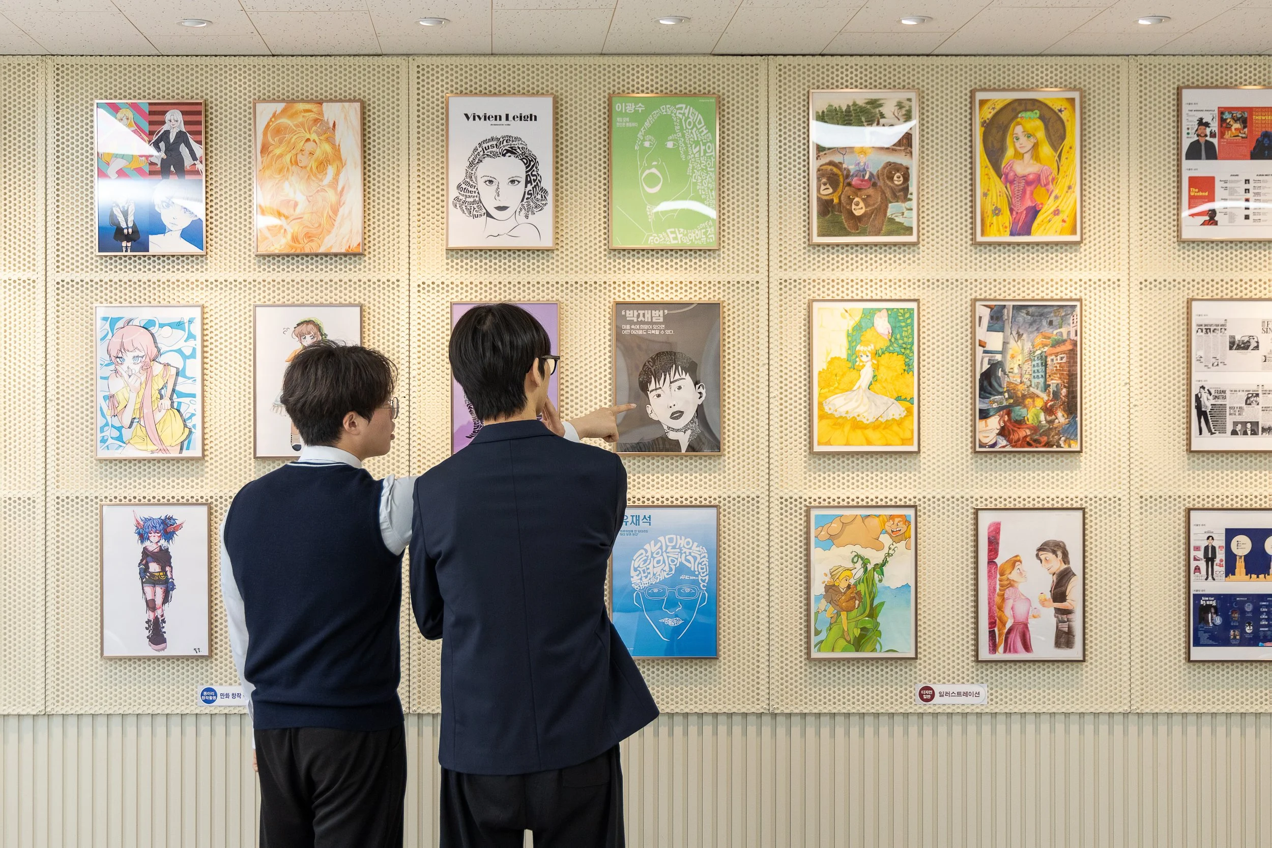 Two young men in black suits and glasses looking at and discussing framed artwork on a beige perforated wall in an art gallery.