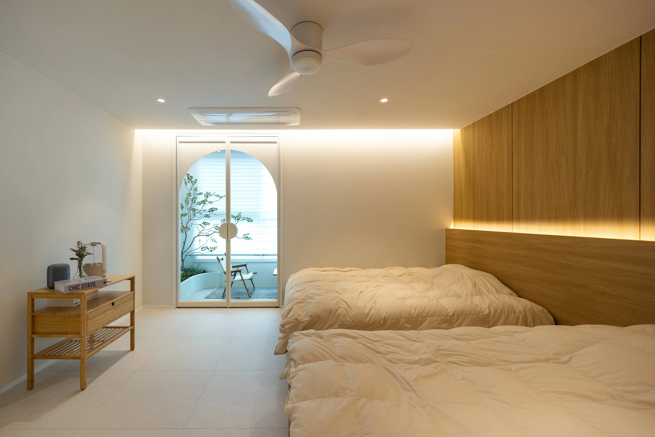 Minimalist bedroom with two beds, wooden headboard, sliding glass door leading to a small balcony with a chair and plant, ceiling fan, and ambient lighting.