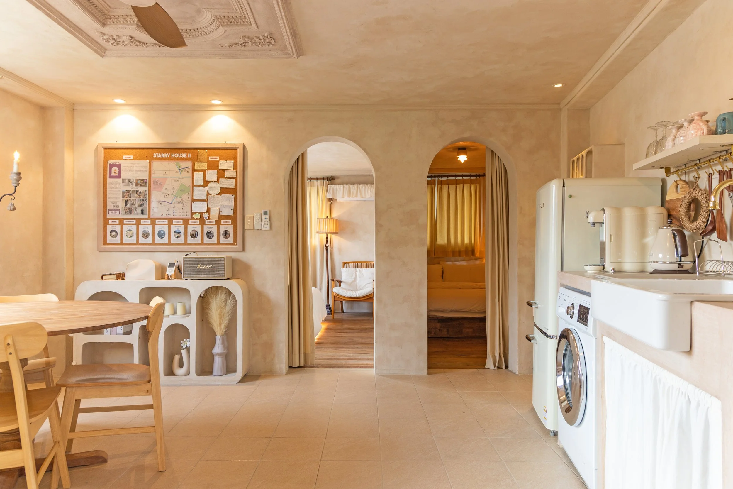 A cozy kitchen and dining area with beige walls and tile flooring, showing a wooden dining table with chairs, a white shelving unit, a vintage fridge, a washing machine, and a countertop with kitchen appliances. There are two doorways leading to bedr