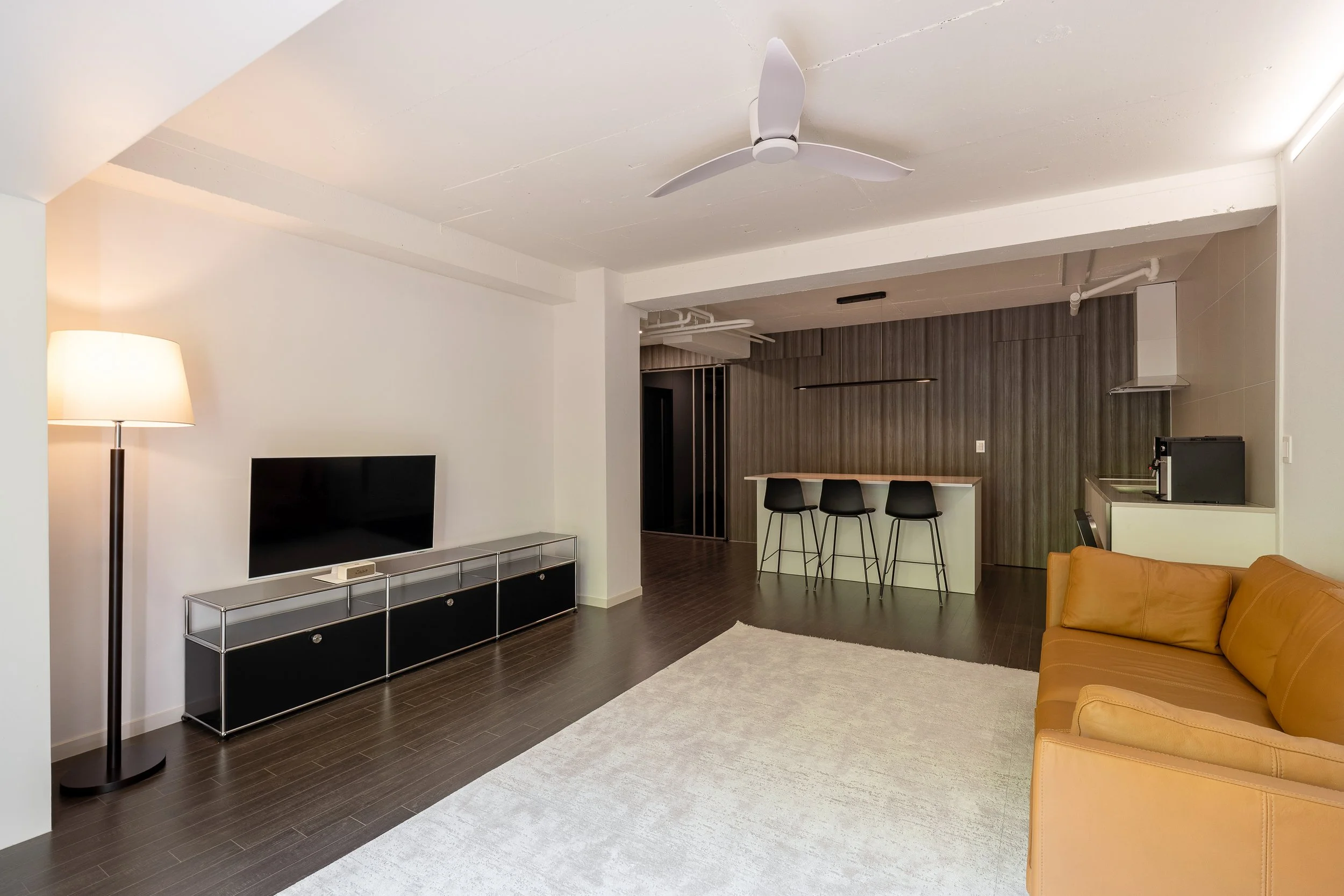Modern living room with a beige sofa, a flat-screen TV on a black and glass console, a tall floor lamp, a white ceiling fan, a dark wood floor, a light-colored rug, and a small kitchenette with a breakfast bar and three black chairs.