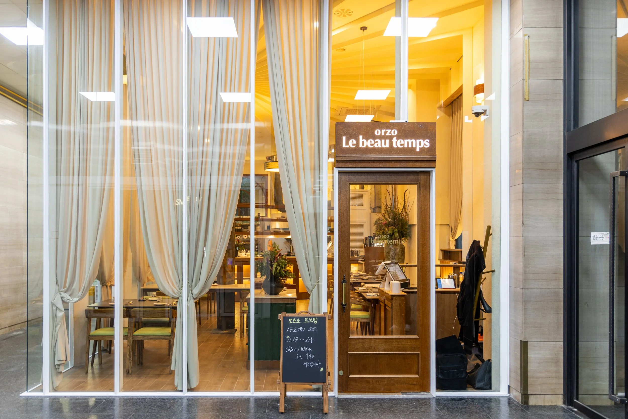 A cozy restaurant with large glass windows and beige curtains, wooden interior, and a sign that reads 'orzo Le beau temps' at the entrance.