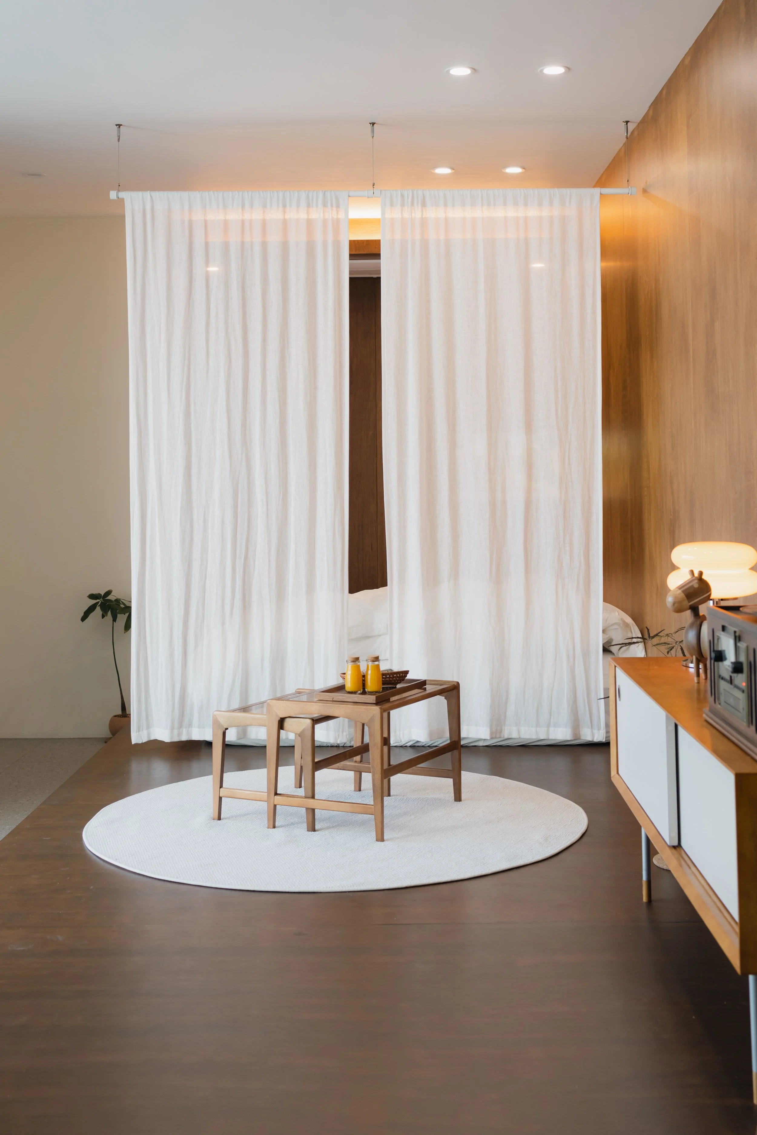 Minimalist room with a white curtain dividing the space, a small wooden table with orange juice bottles, a round white rug, and a wooden sideboard with a lamp and stereo system.