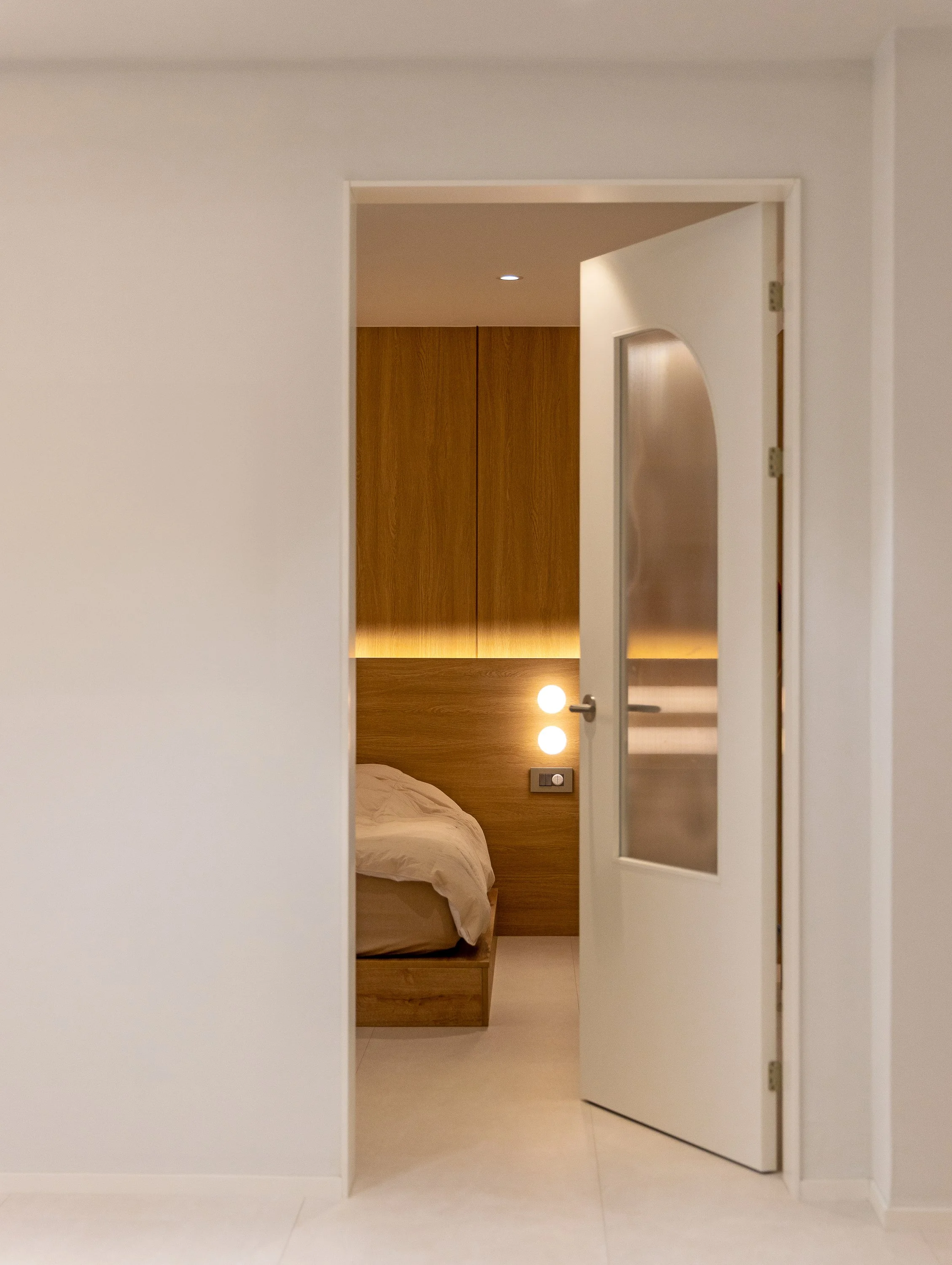 View through an open door into a bedroom with a wooden headboard, soft beige bedding, and modern wall-mounted lamps.