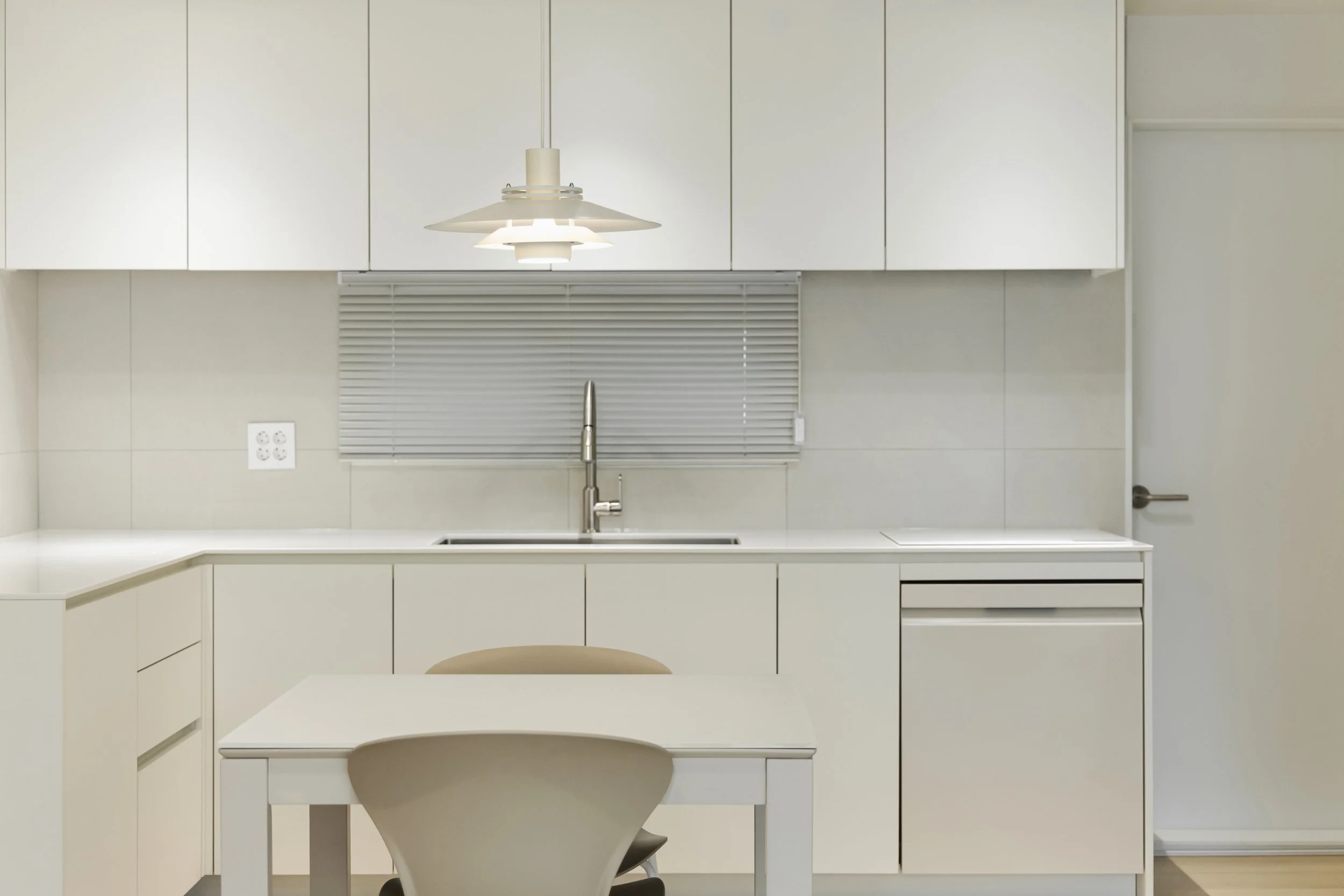 Minimalist kitchen with white cabinets, a stainless steel sink, a modern hanging light fixture, closed blinds, and a white dining table with chairs.