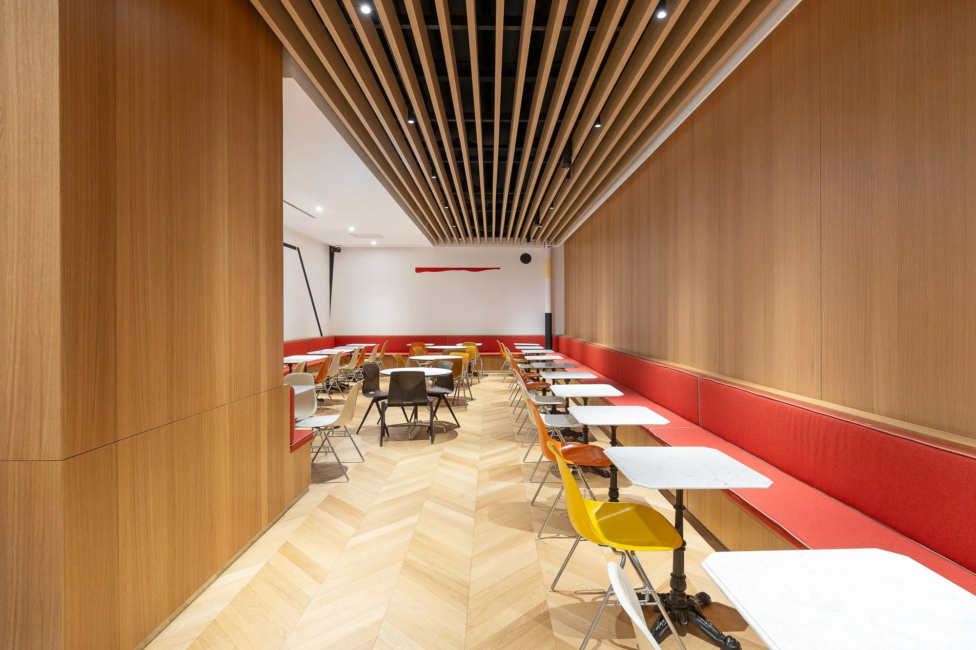 Empty modern cafe with wooden wall and ceiling design, white marble tables, colorful chairs, and red cushioned bench along the wall