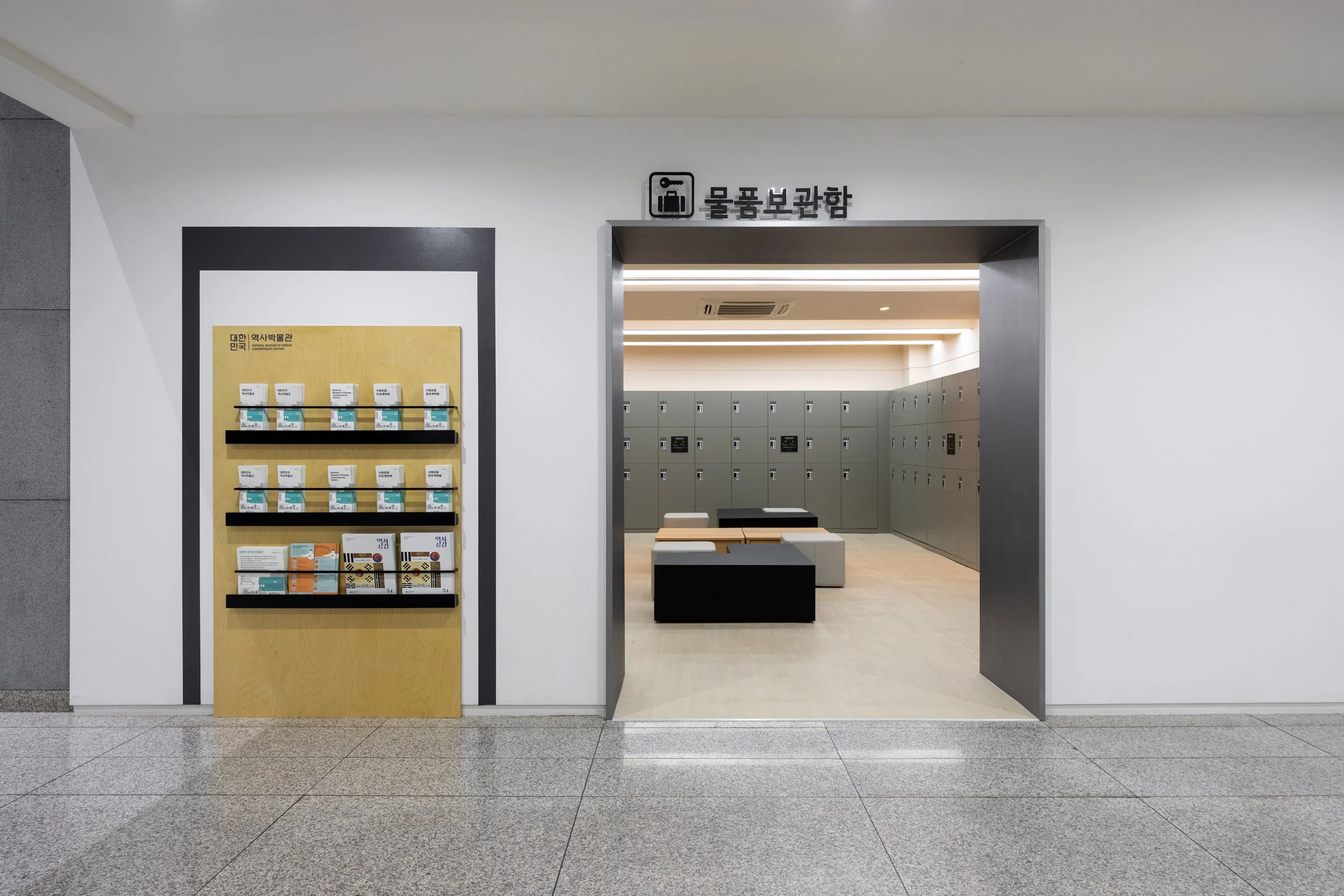 Entrance to a locker room with gray lockers and wooden benches, sign in Korean indicating a lost and found area, informational brochure display outside.