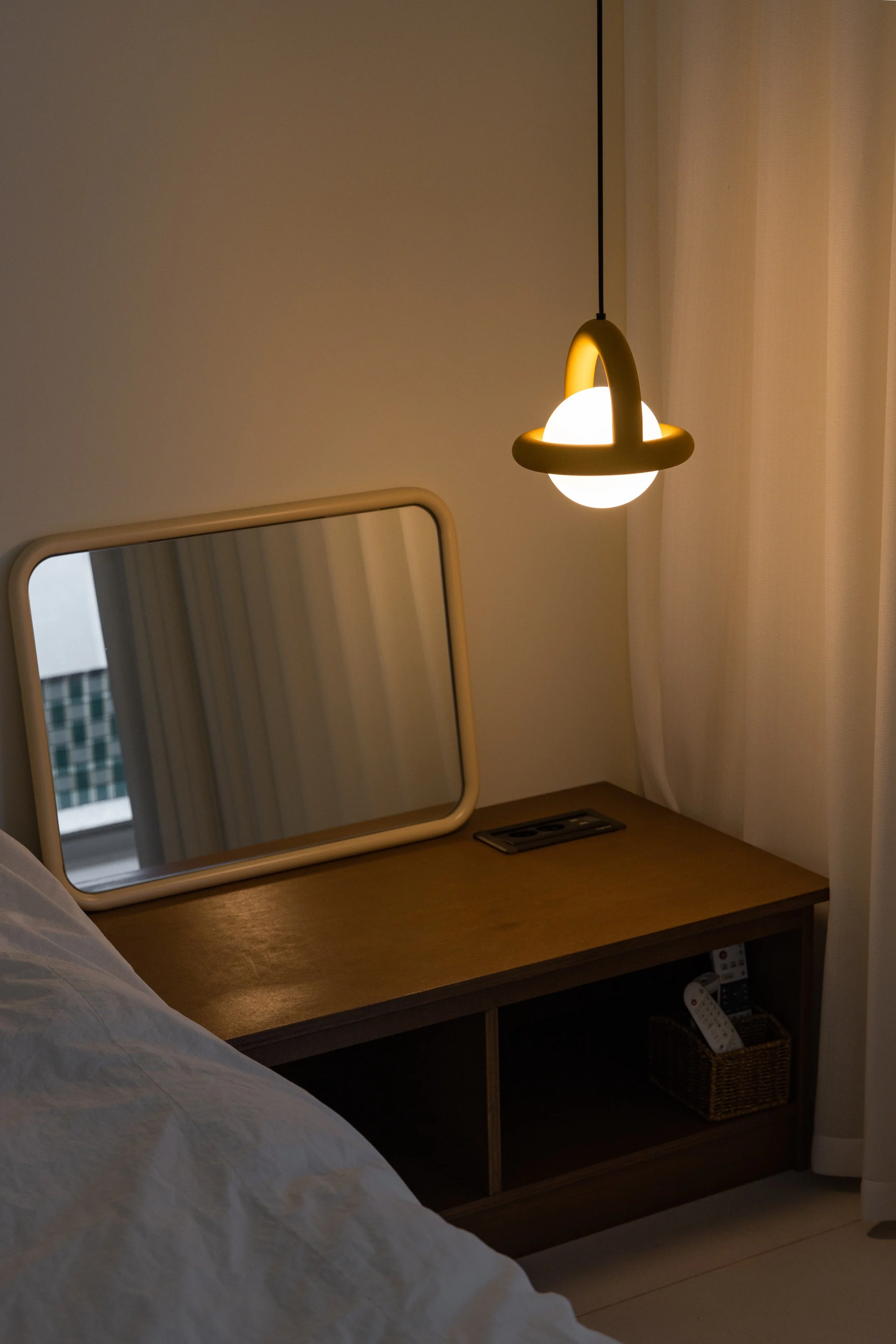 A hotel bedside table with a mirror, a remote control, a woven basket with remote controls, and a hanging lamp with a round light bulb.