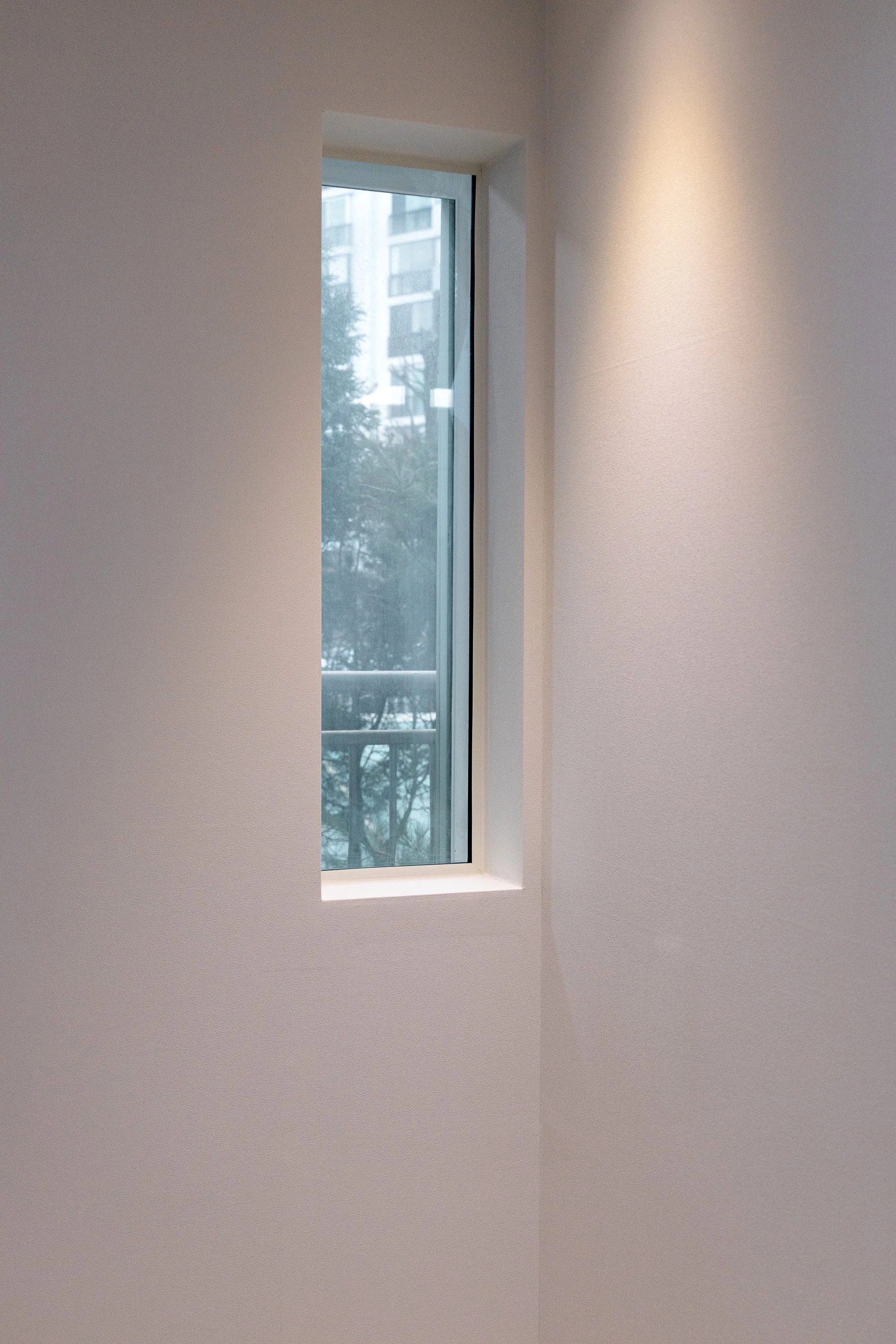 An interior view of a room with a tall, narrow window on a plain white wall, showing a cityscape with buildings and trees outside.