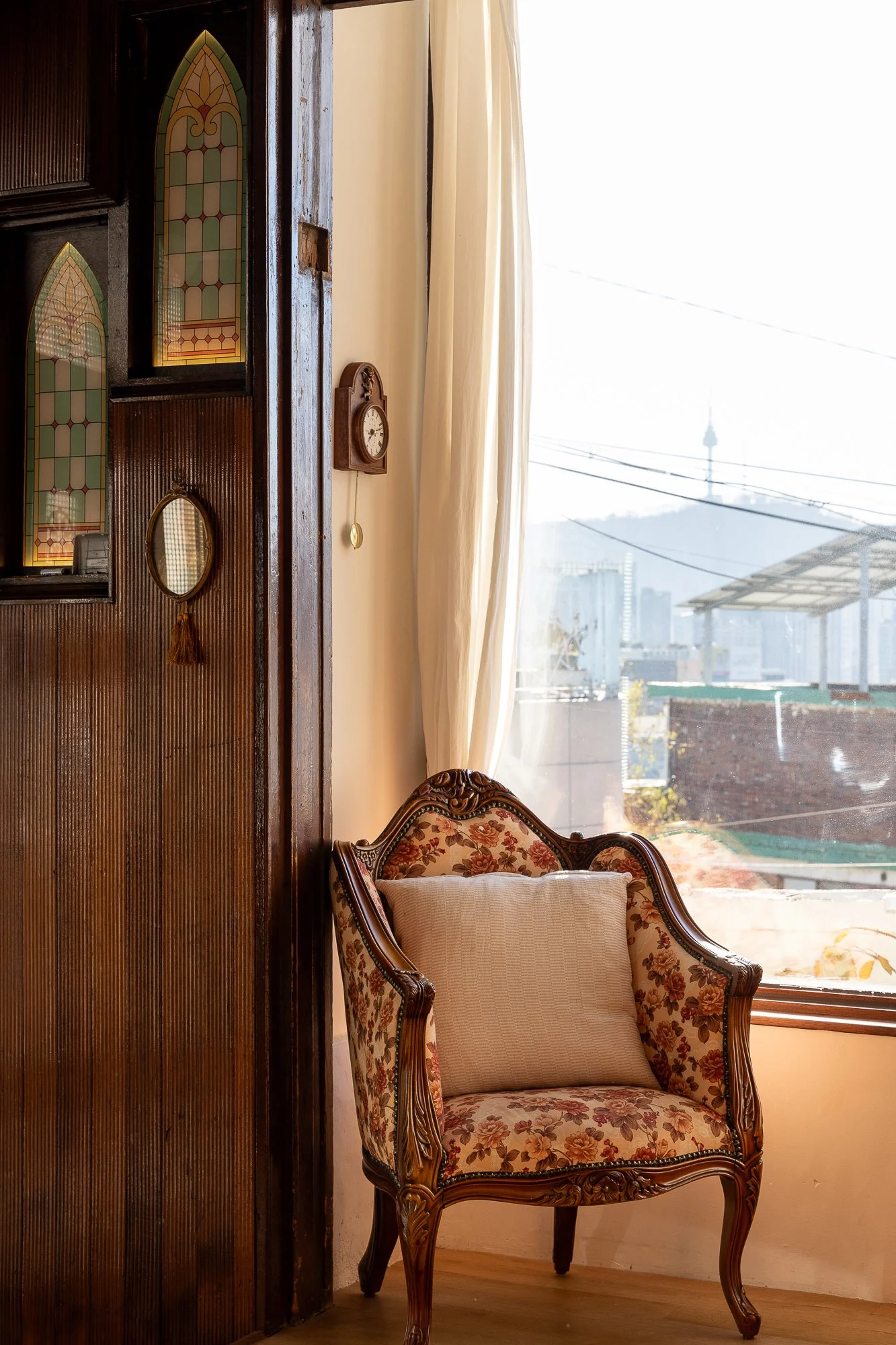 An indoor scene with an ornate floral armchair next to a window with white curtains, a small clock on the wall, and stained glass decorations on a wooden wall. Outside the window, city buildings and a tower are visible.