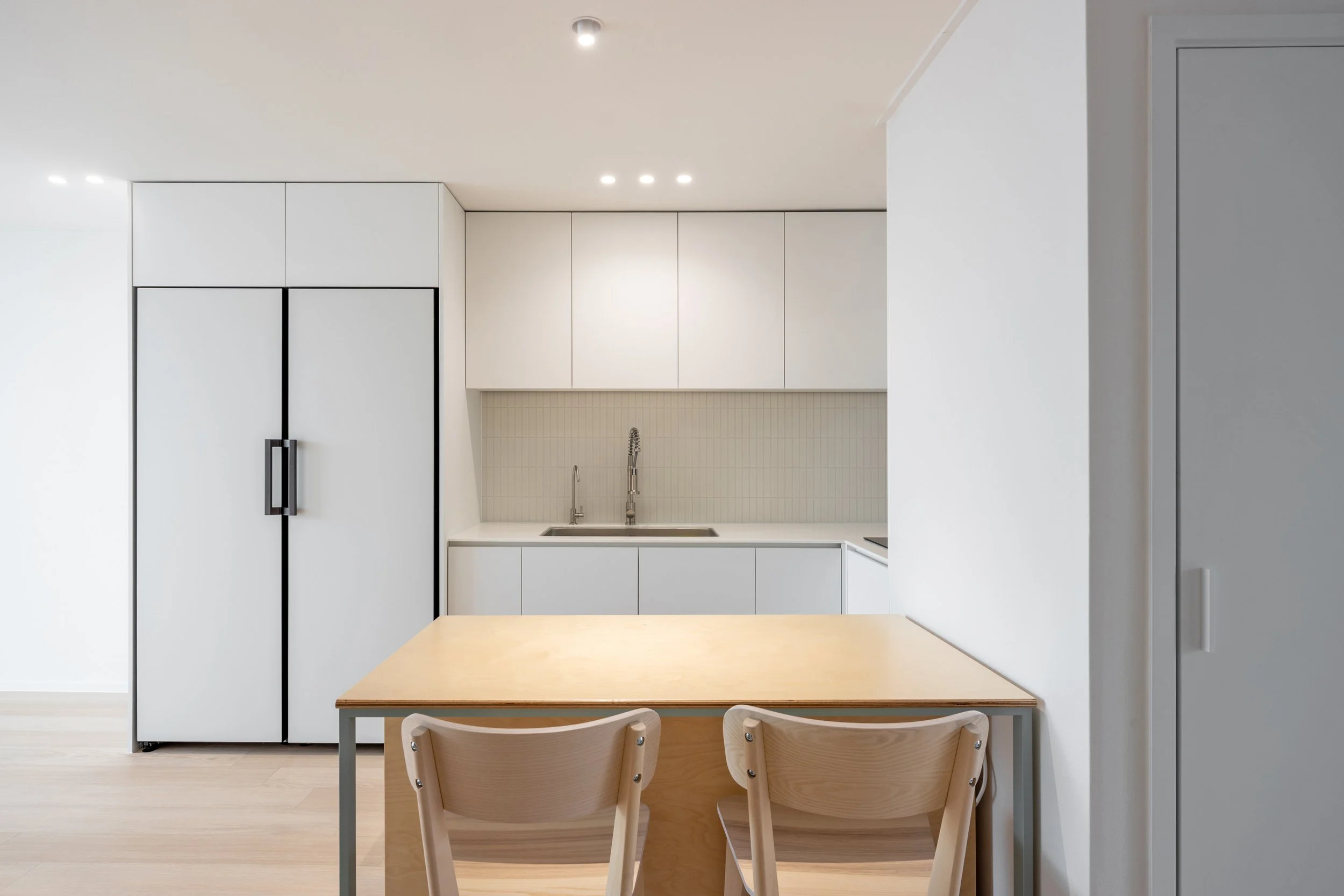 Minimalist kitchen with white cabinets, a small wooden dining table, two wooden chairs, and built-in appliances.