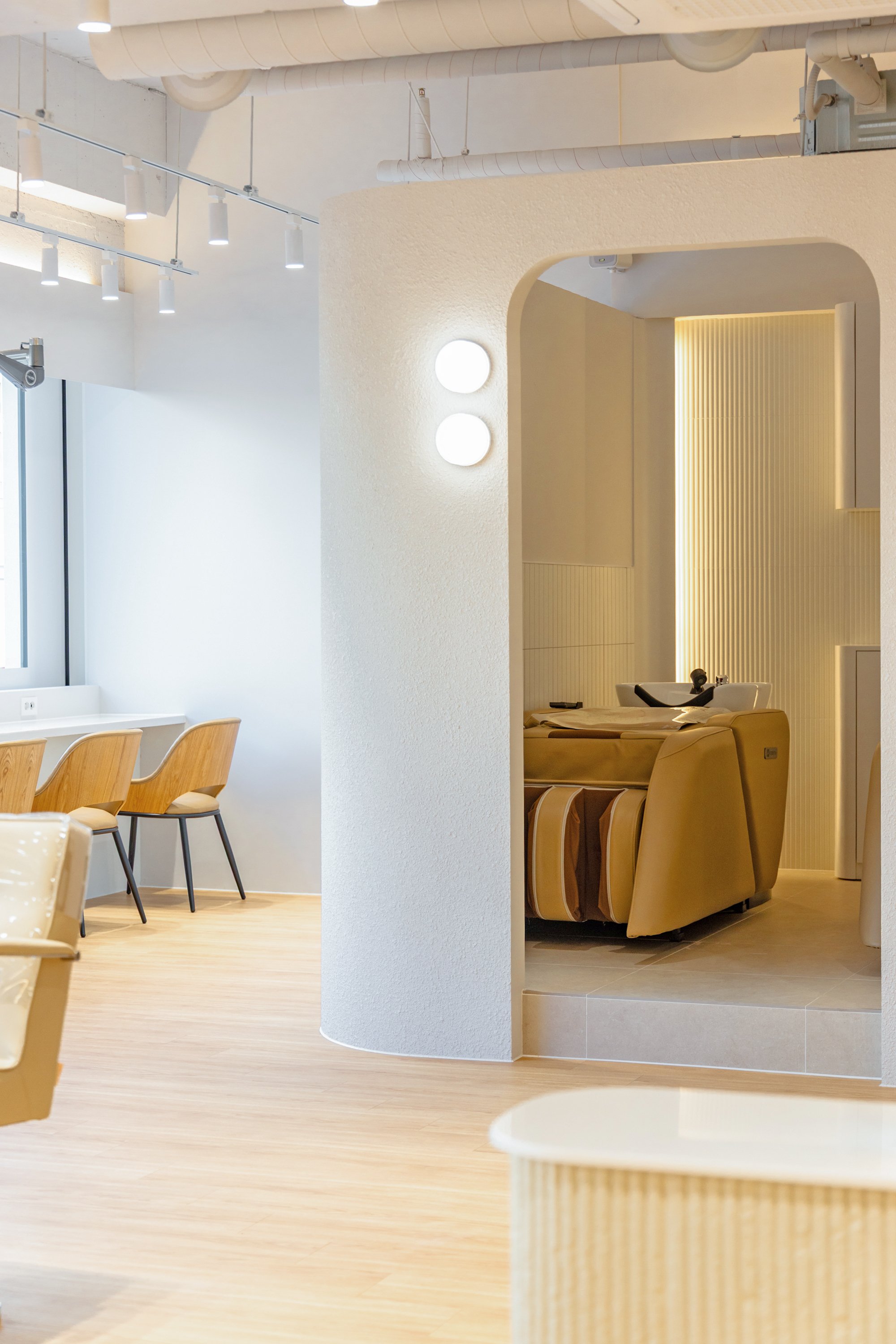 Modern salon with white walls, wooden chairs, and a beige shampoo station.
