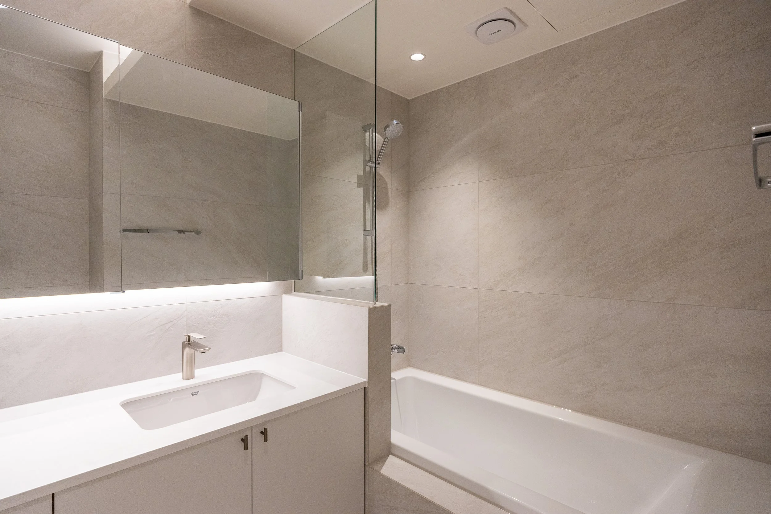 Modern bathroom with beige tiled walls, a white vanity with a sink and faucet, a large mirror, a bathtub, and a walk-in shower with a glass partition, illuminated by ceiling lights.