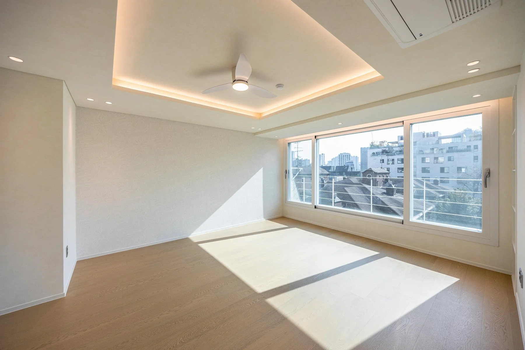 Empty room with large windows, wooden floor, ceiling fan, and recessed lighting, cityscape outside.