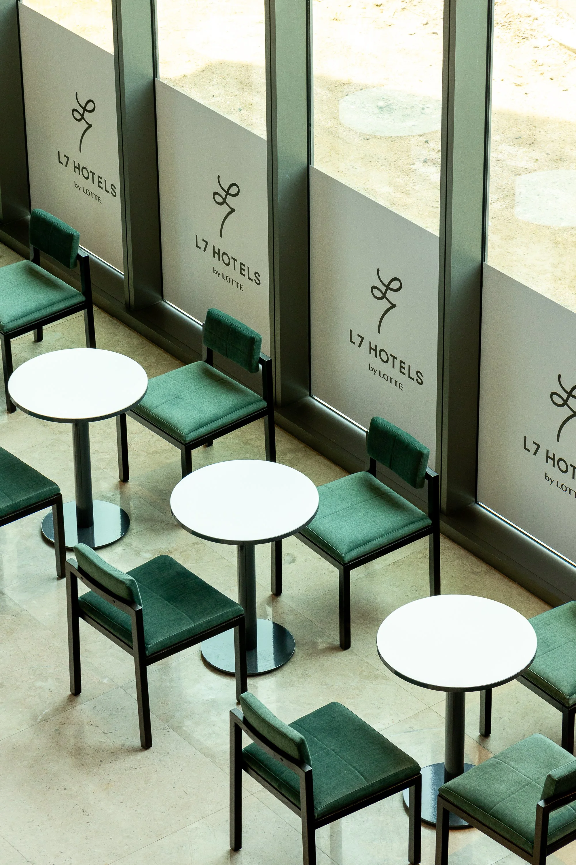 Empty seating area with green chairs and white round tables near large windows with posters for L7 Hotels by Lotte.