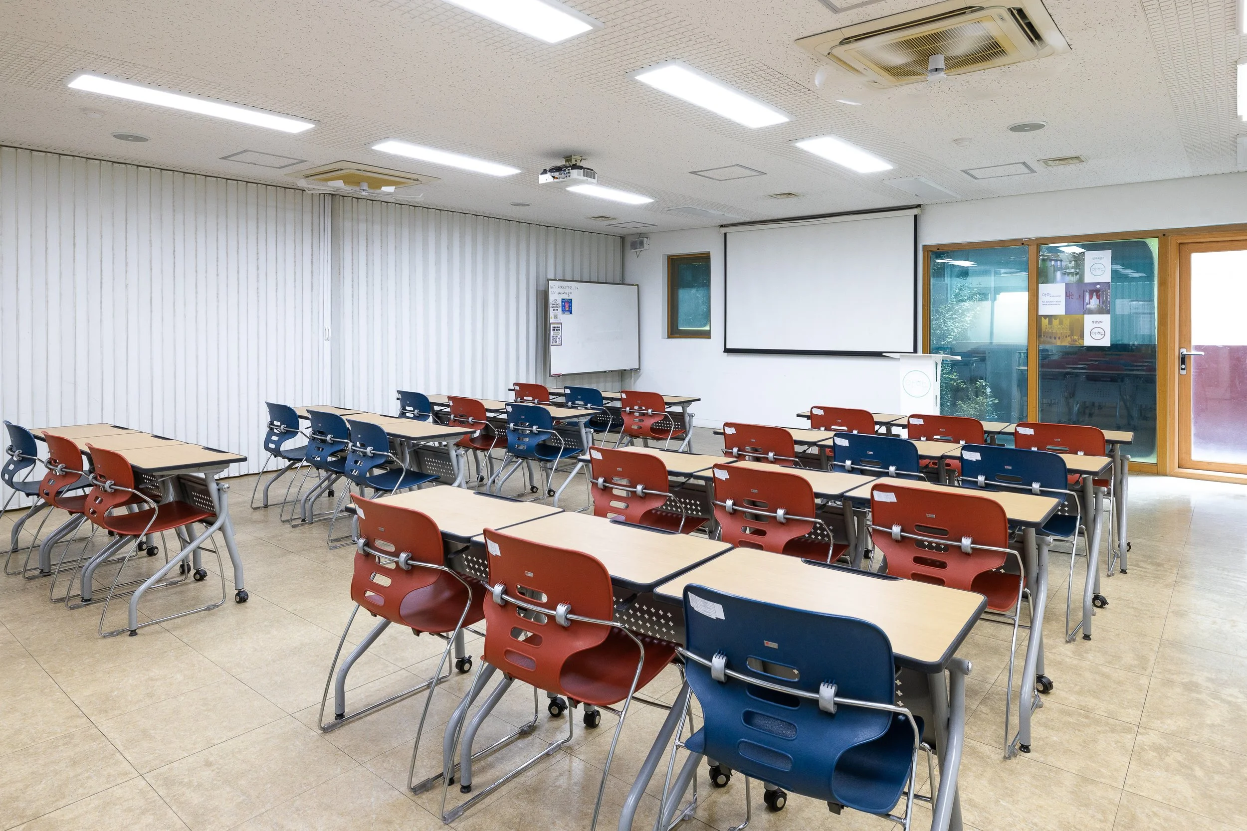 Empty classroom with rows of school desks and chairs, whiteboard, projector screen, and a door with glass window.