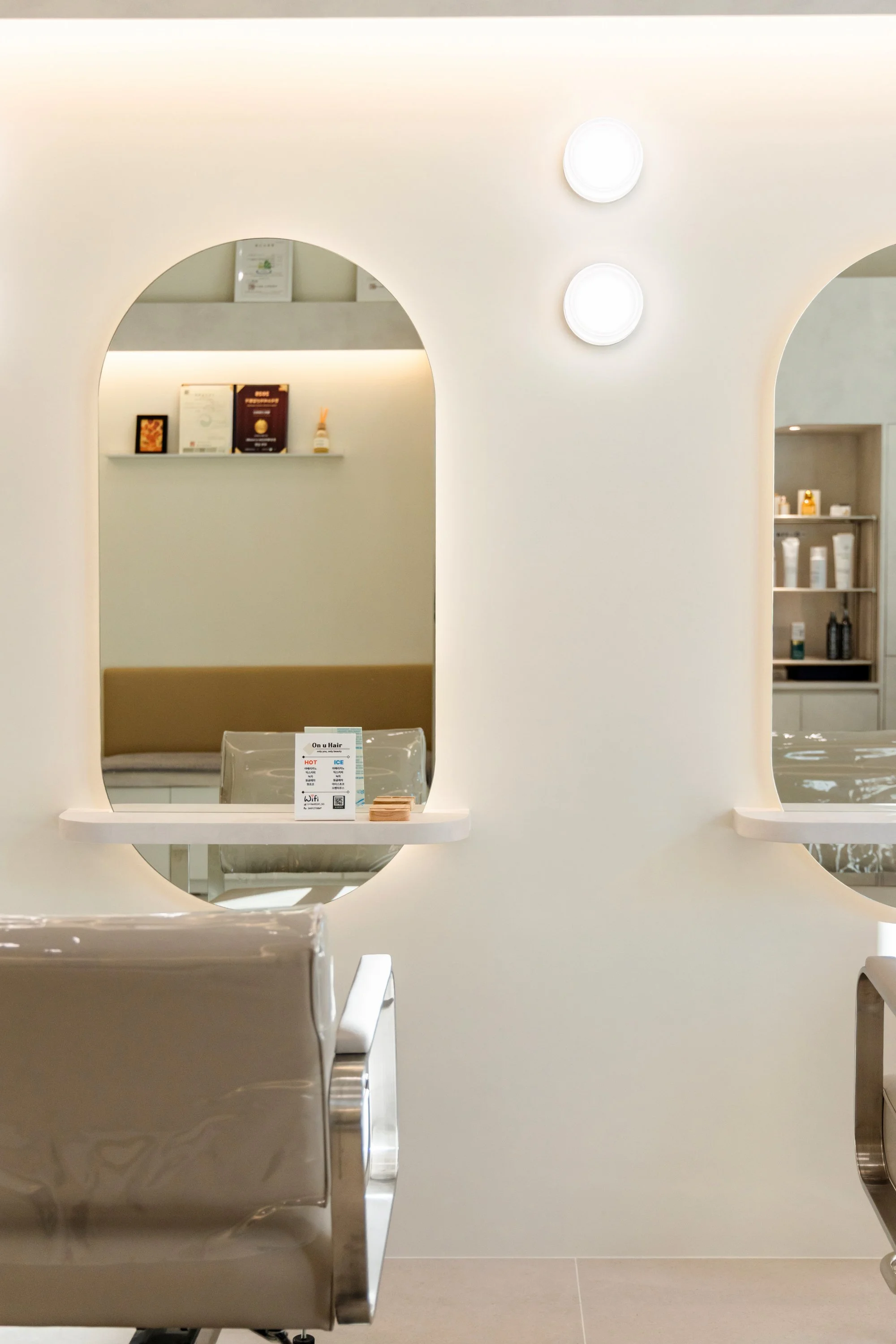 Interior of a modern, minimalist salon or spa with white arched wall mirrors, beige chairs, and shelves with beauty products.