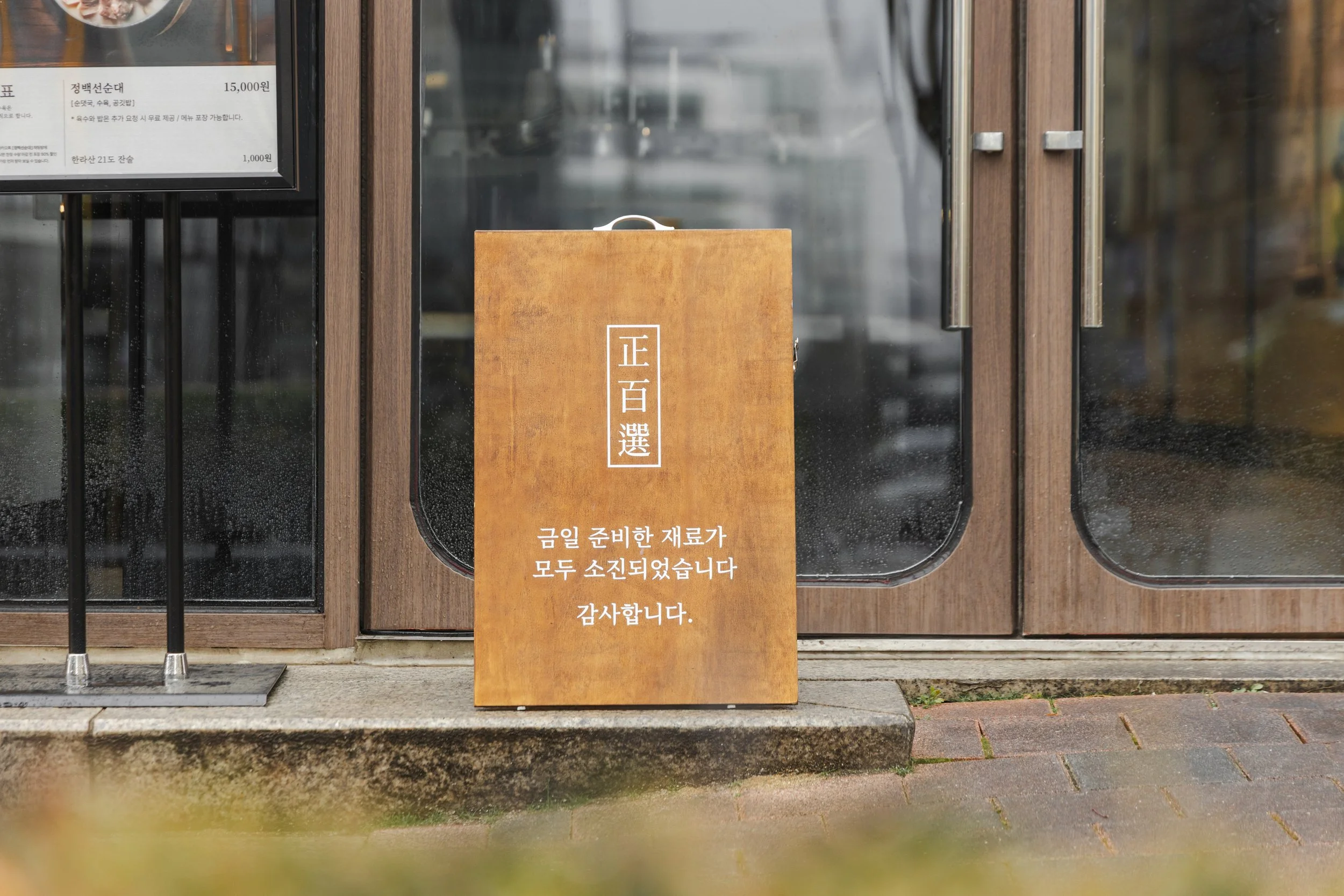 Wooden sign in front of a glass door with Korean writing, indicating the ingredients for the day are all sold out.