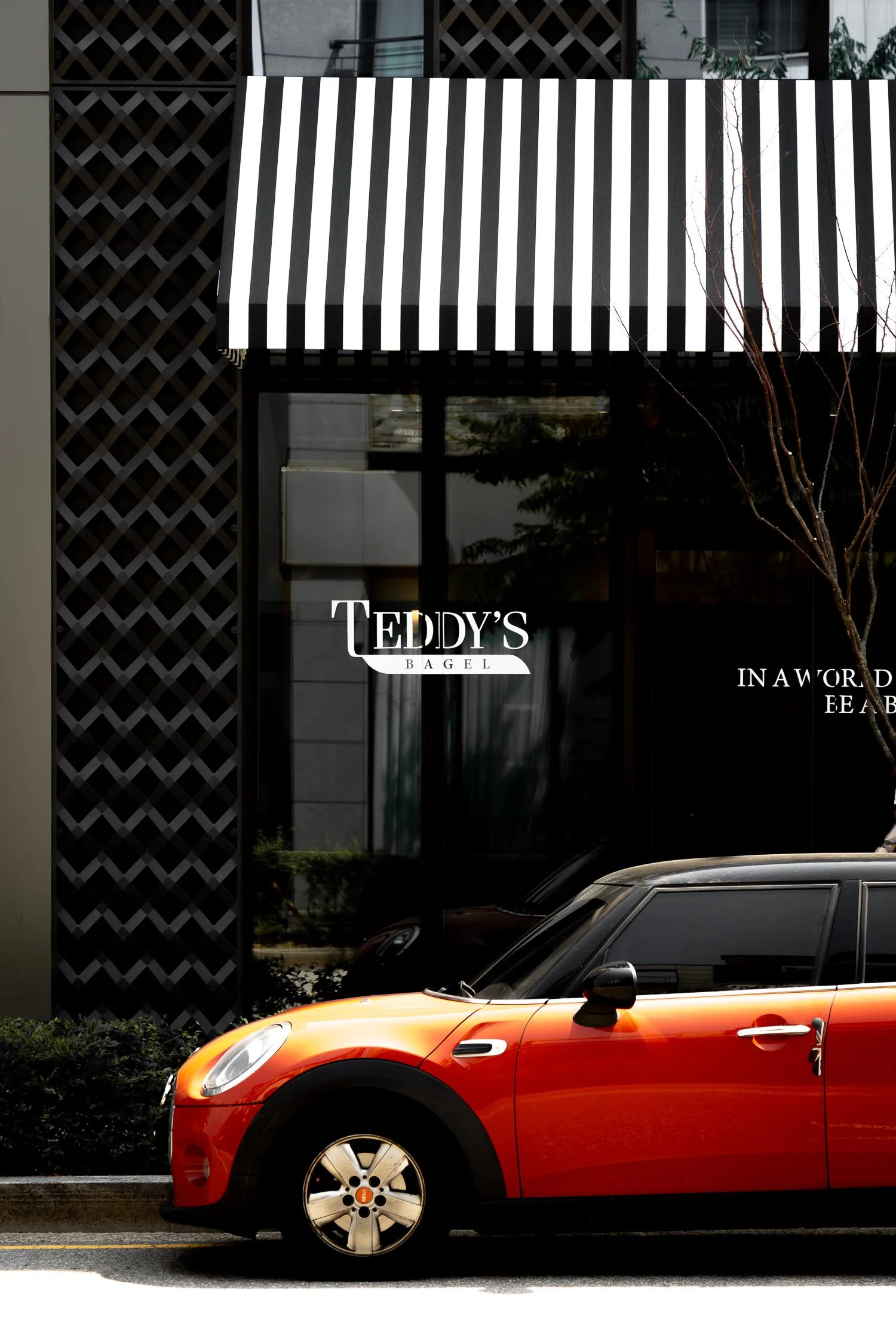 Orange and black Mini Cooper parked in front of a glass storefront with a striped black and white awning. The store sign reads "Teddy's Bagel."