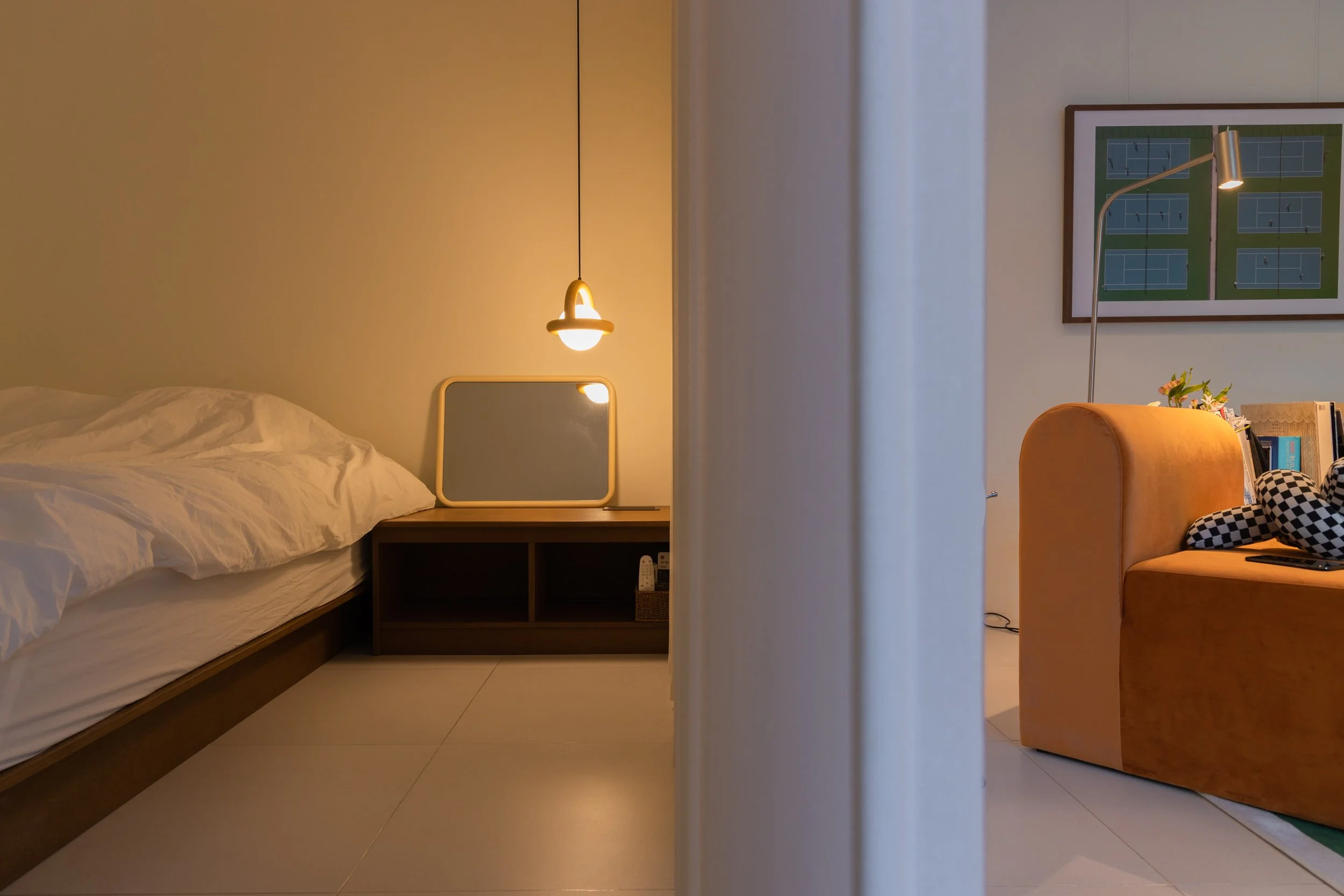 A split view of two rooms separated by a curtain. The left side shows a bedroom with a bed, a mirror on a bedside table, and a hanging lamp. The right side features a living room with an orange sofa, a floor lamp, a framed picture of tennis courts, a
