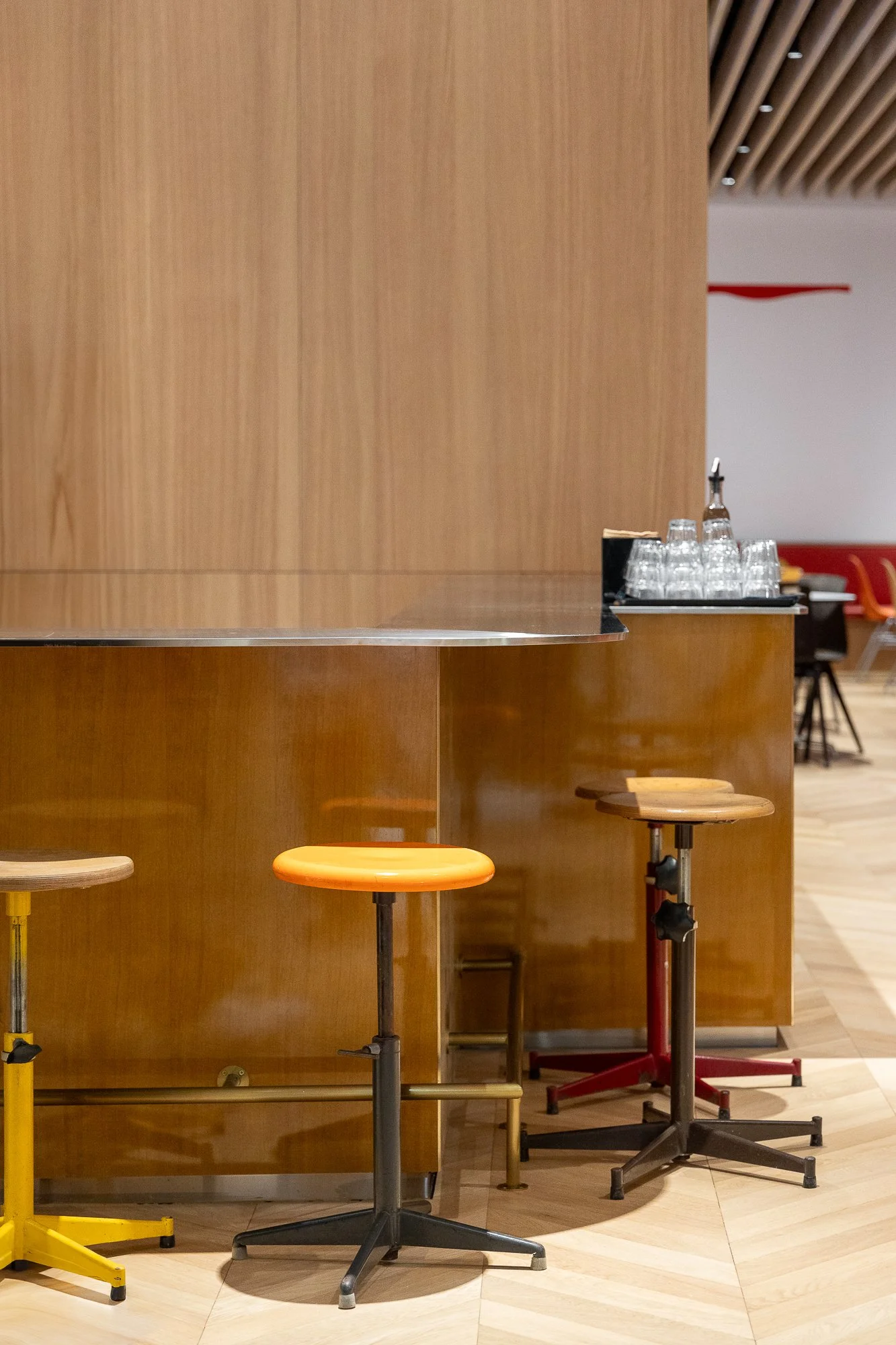 View of a cafeteria with wood-paneled counters, bar stools in orange, yellow, and black, and a tray of glasses and water bottles on the counter, with tables and chairs in the background.