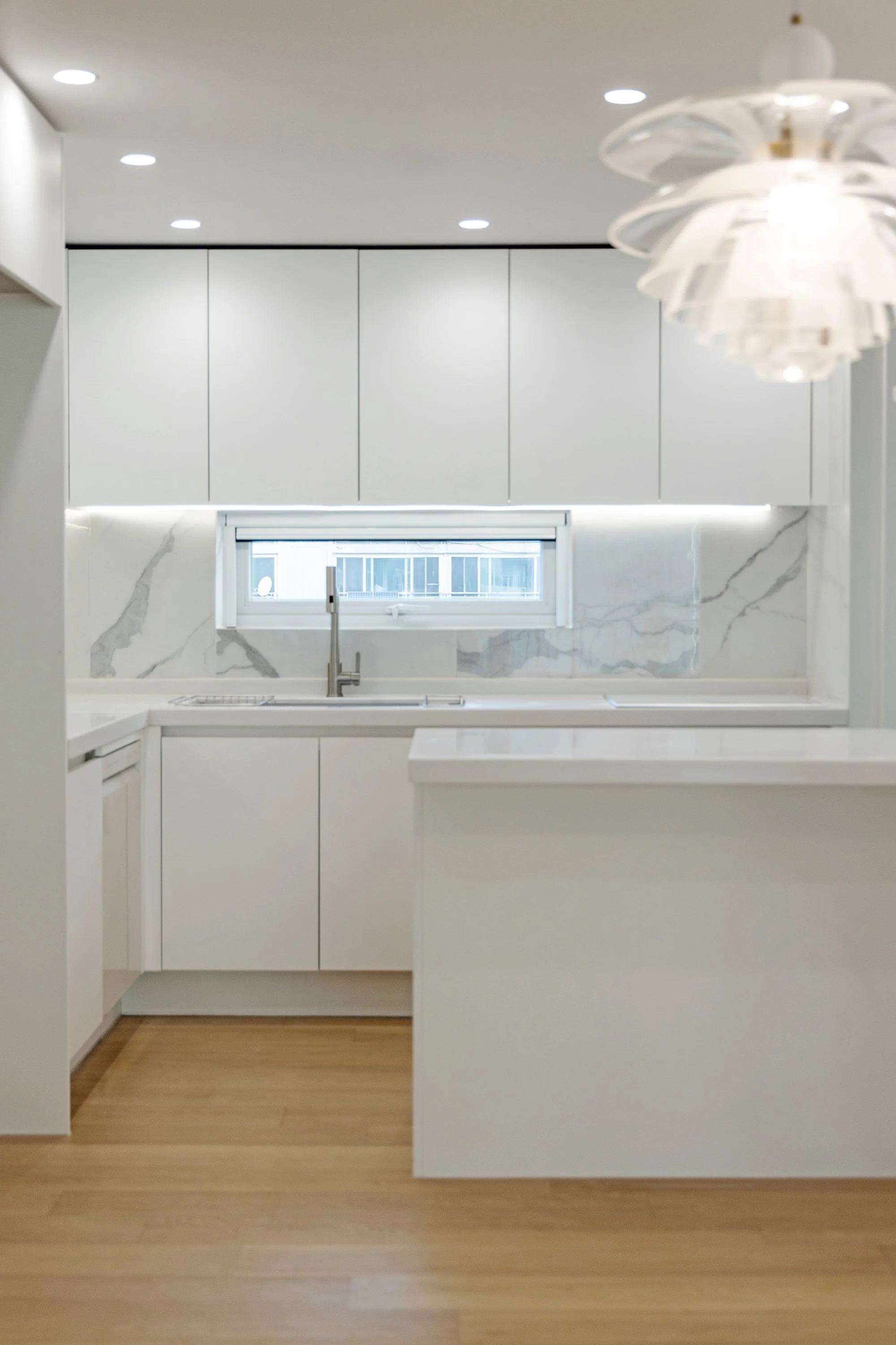 Modern white kitchen with marble backsplash, window above sink, wooden floor, and ceiling lights.