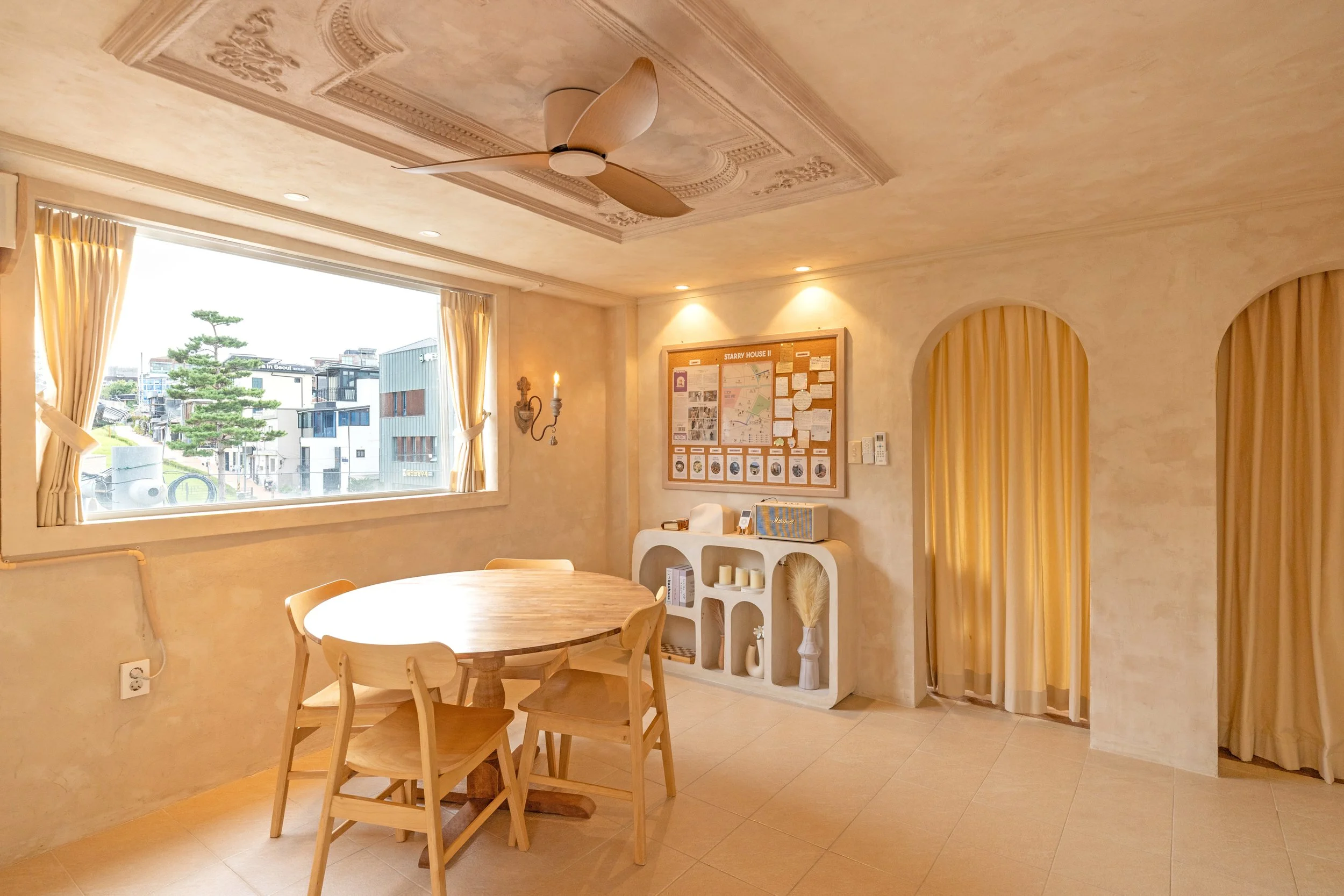 A cozy dining area with a round wooden table and four matching chairs, a large window with beige curtains showing a city view, a bulletin board on the wall, and decorative shelving units filled with candles and ornaments; warm lighting enhances the i